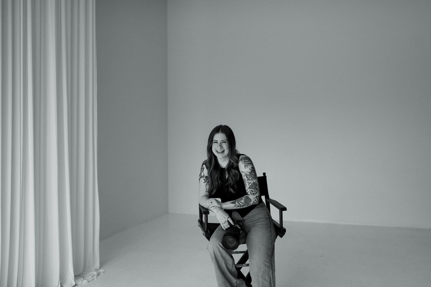 Black and white photo of tattooed female photographer sitting in director chair smiling in bright studio.