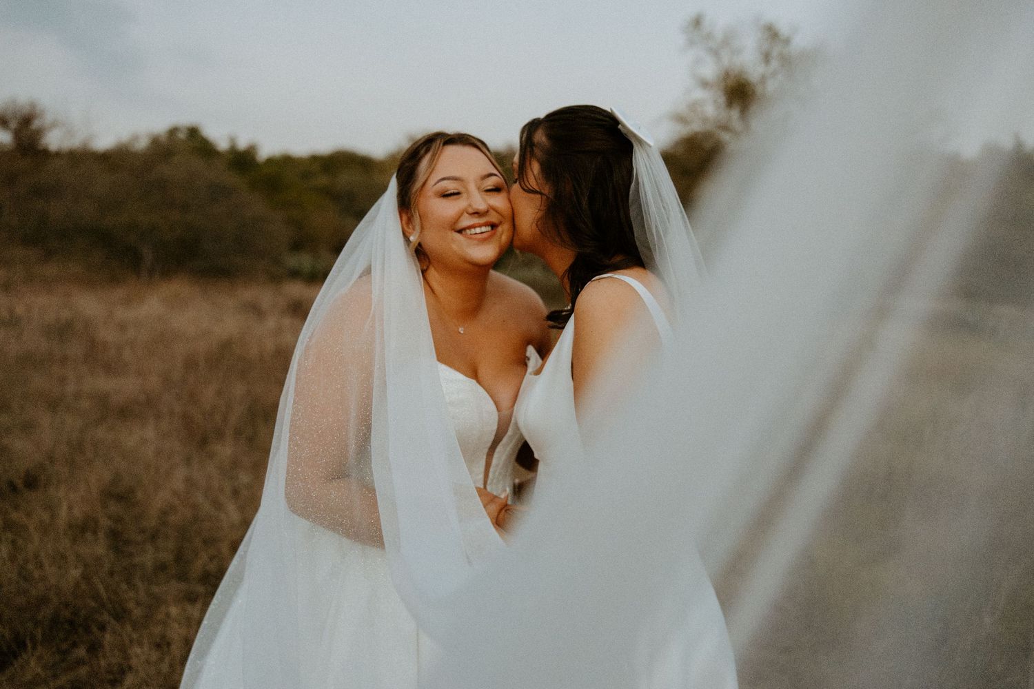 Two brides in white wedding gowns share a tender kiss outdoors in golden hour light with flowing veil.