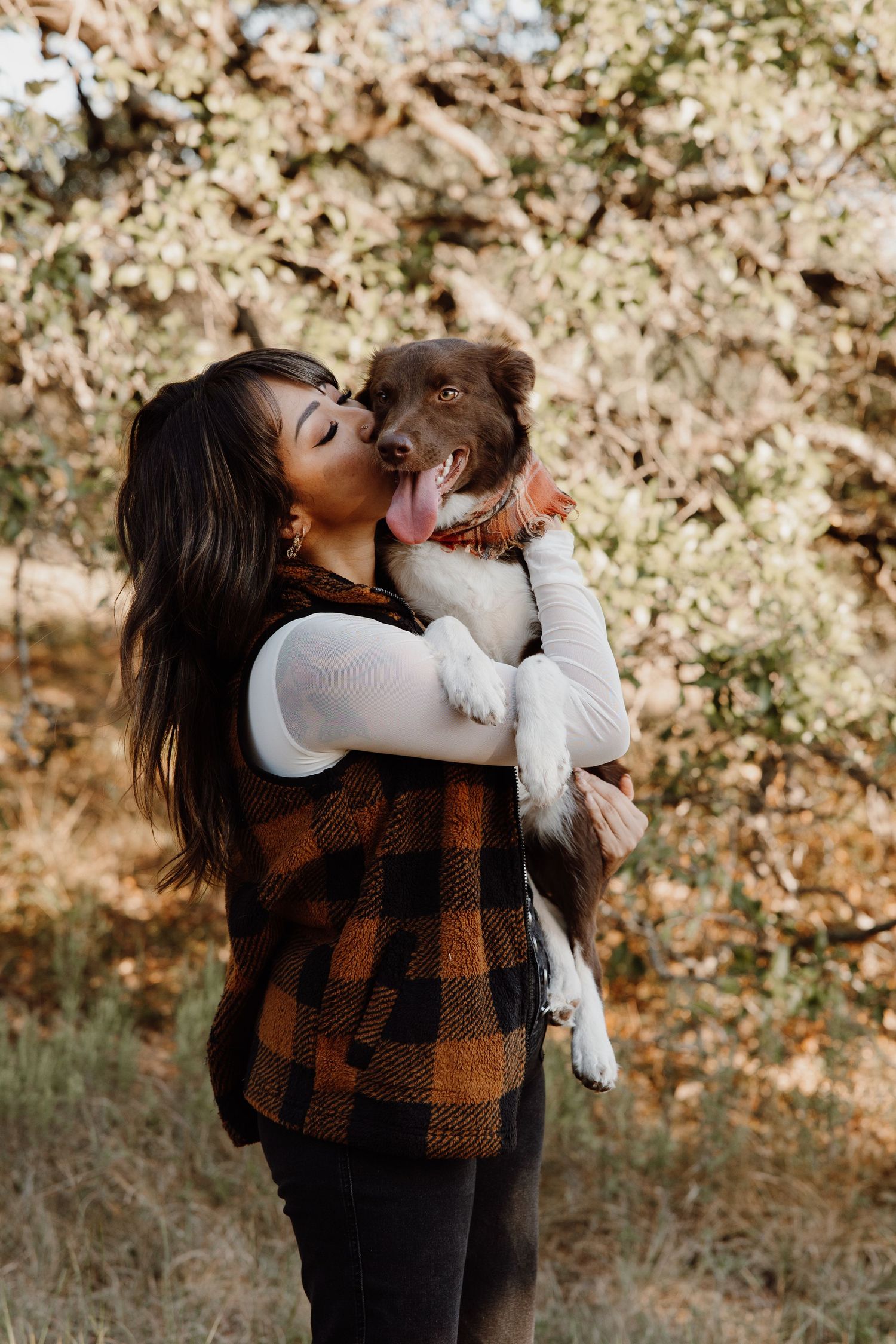 Woman kissing a small brown and white dog she is holding in an autumn outdoor setting.