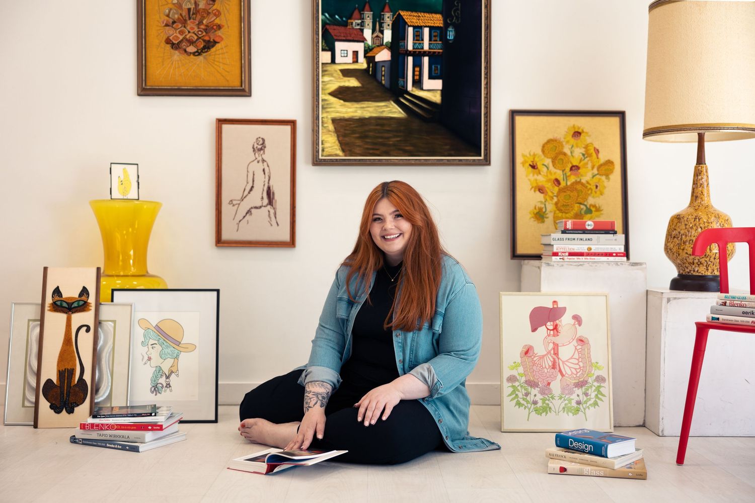 Redheaded woman sitting on floor reviewing artwork surrounded by vintage paintings in white room.