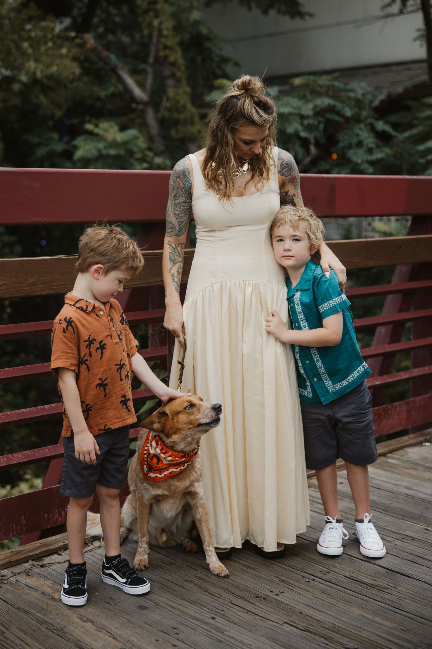 A tattooed mother in a cream dress stands with her two sons and golden dog on a wooden bridge.