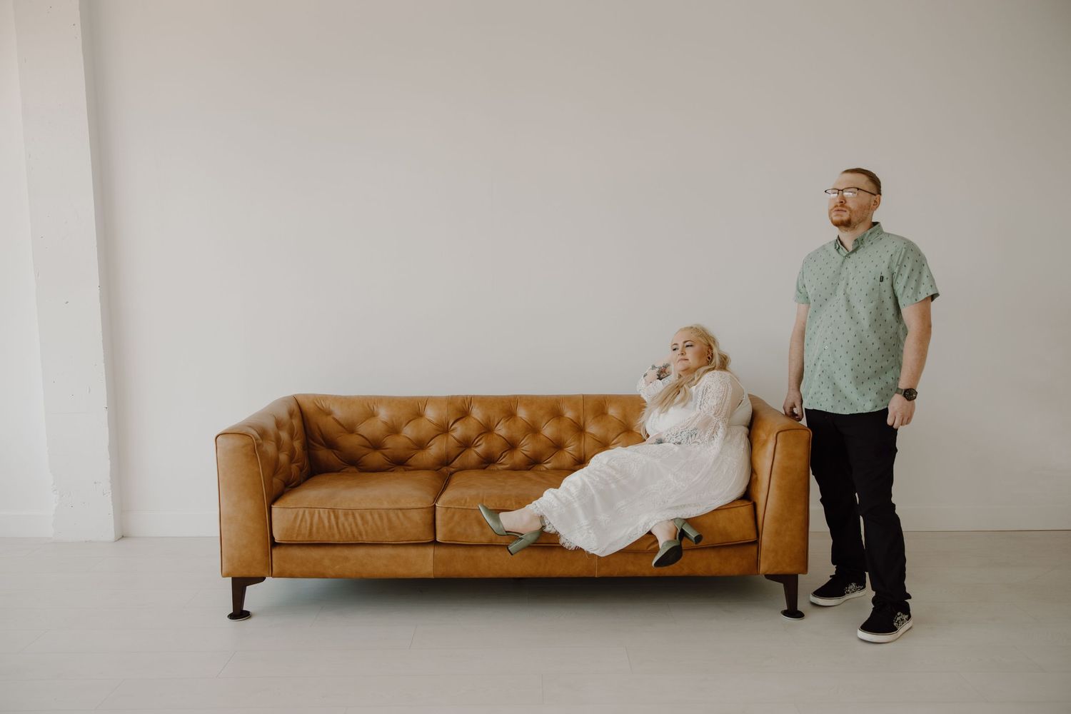 Bride relaxing on tan leather sofa while groom stands beside her in bright white studio.