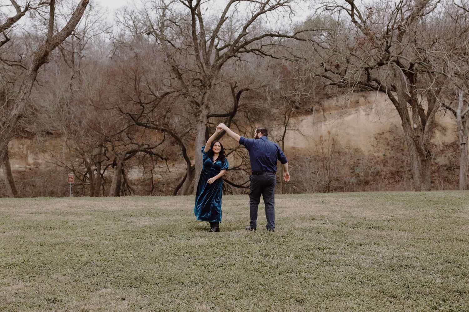 Couple dancing and spinning joyfully in grassy field surrounded by bare winter trees.