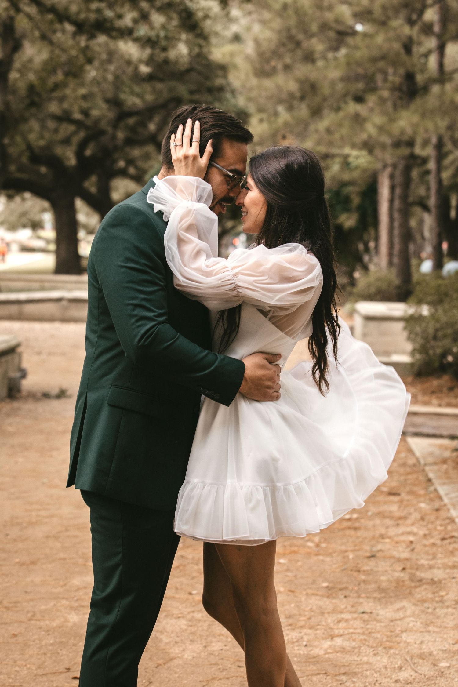 Bride and groom share a romantic moment about to kiss outdoors, she in white mini dress, he in dark green suit.
