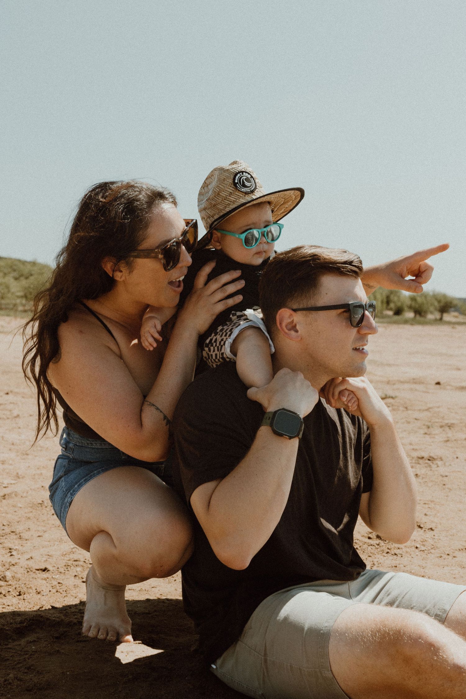 Parents with sunglasses crouching with baby on dad's shoulders pointing into the distance on a sandy beach.