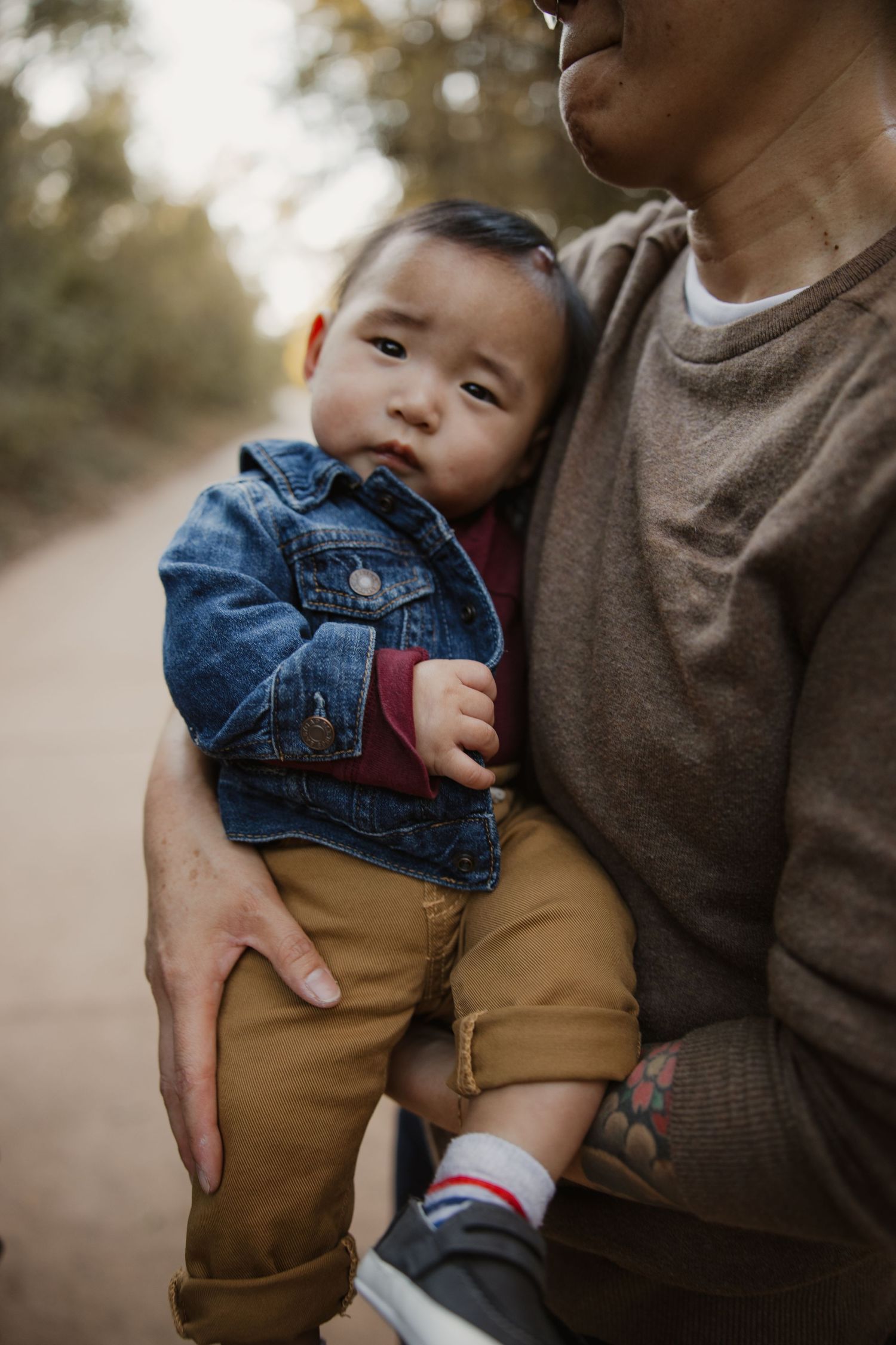 Parent with tattoos holding baby boy in denim jacket and tan pants on an autumn dirt path.