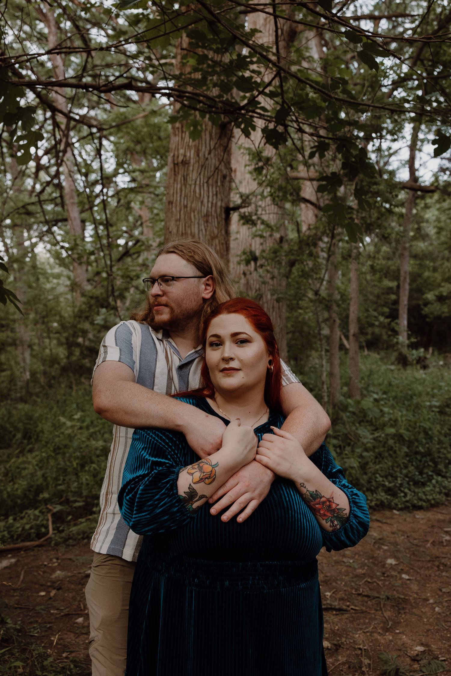 A couple embraces in a lush green forest, the man wrapping his tattooed arms around the woman in a teal velvet dress.