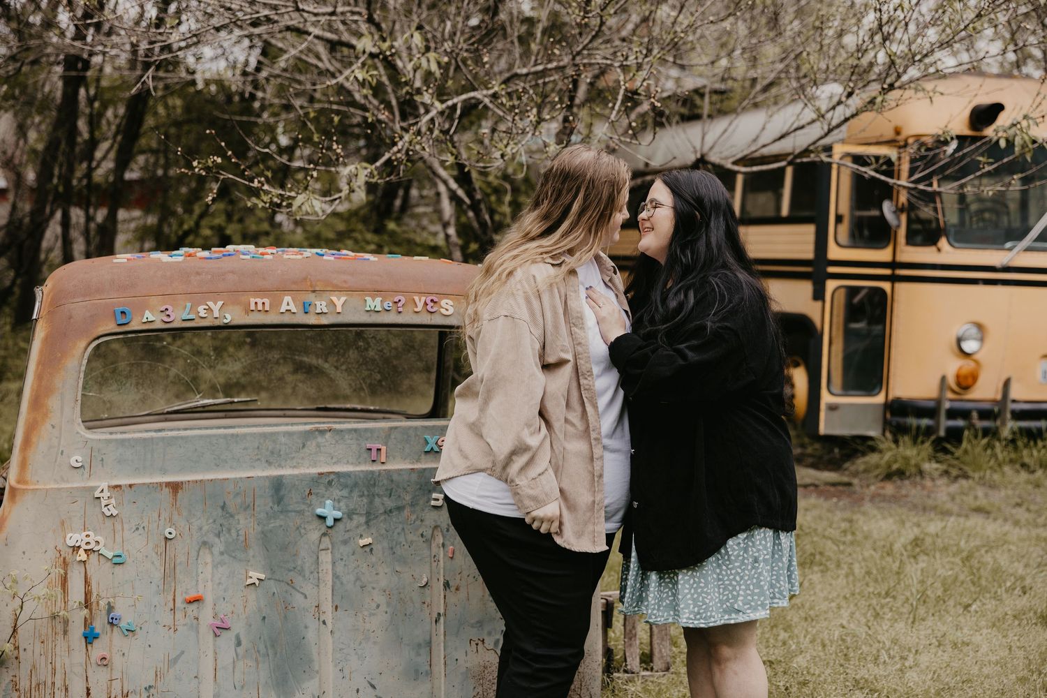 Two women hold hands and share a tender moment beside a rustic vintage truck with a yellow school bus in the background.