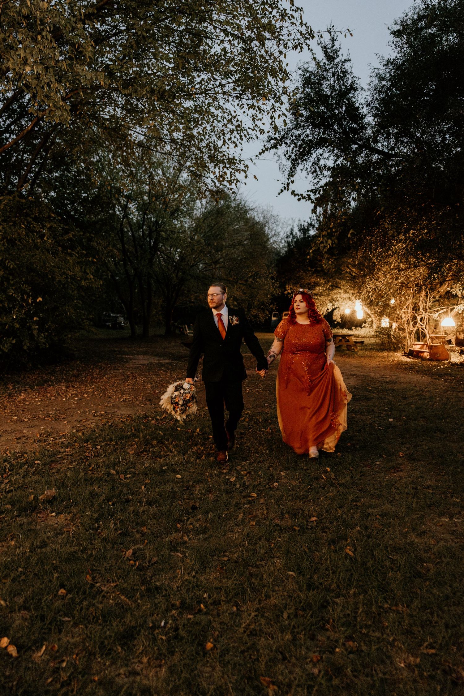 Newlyweds walk hand in hand through autumn garden at twilight holding bridal bouquet.