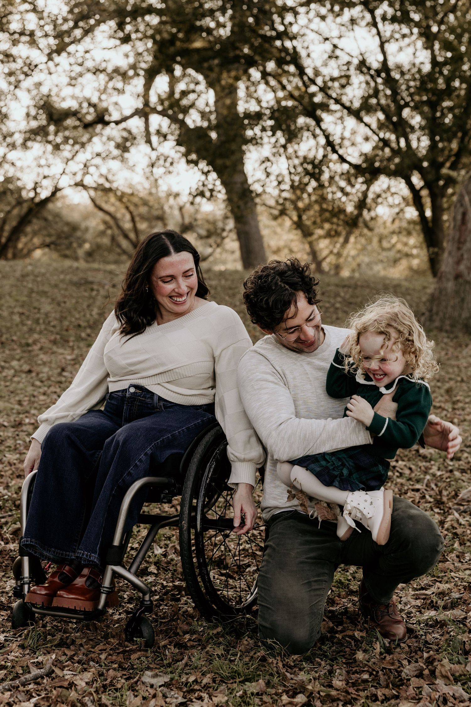 Happy family with mother in wheelchair and father holding toddler daughter laughing together in autumn park setting.