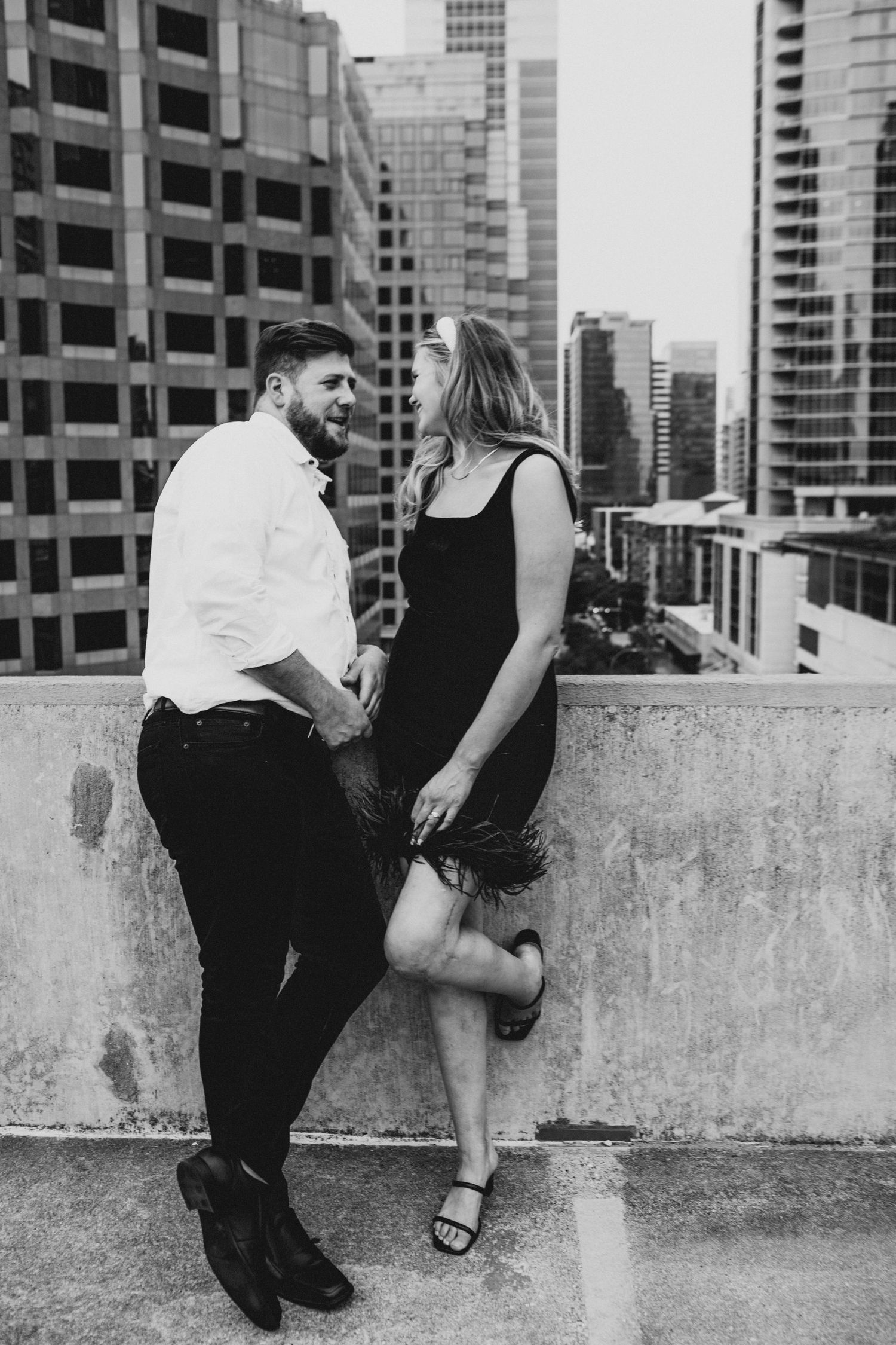 Black and white photo of couple holding hands and laughing on rooftop with city skyline view.