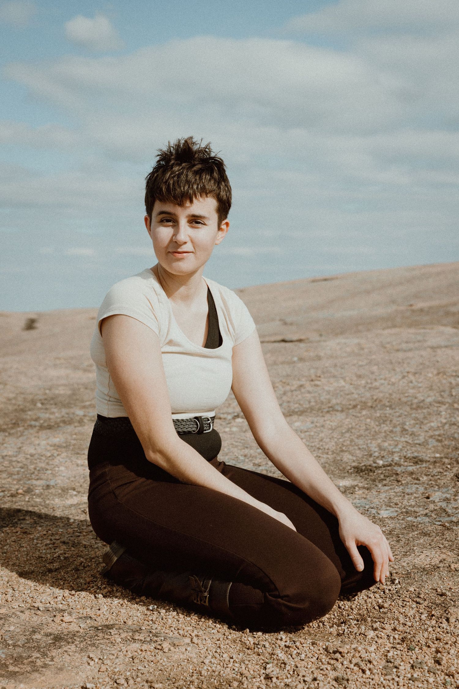 Person with short hair in white crop top and dark pants sitting on rocky terrain.