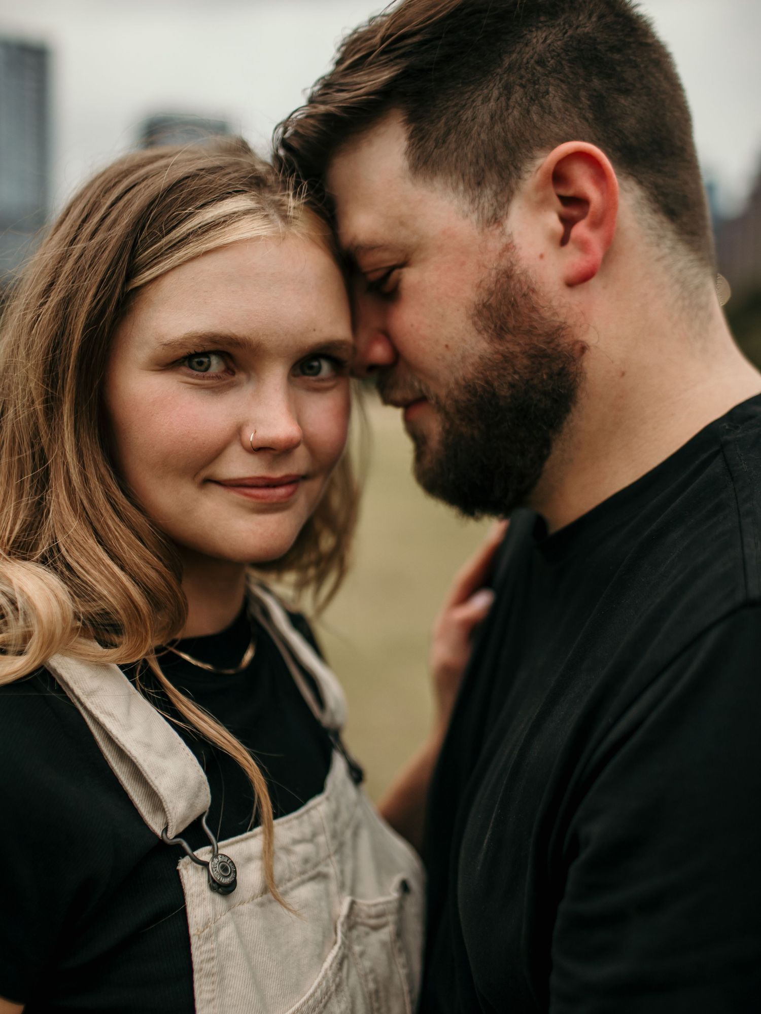 Intimate engagement portrait of bearded man and blonde woman in overalls touching foreheads outdoors.