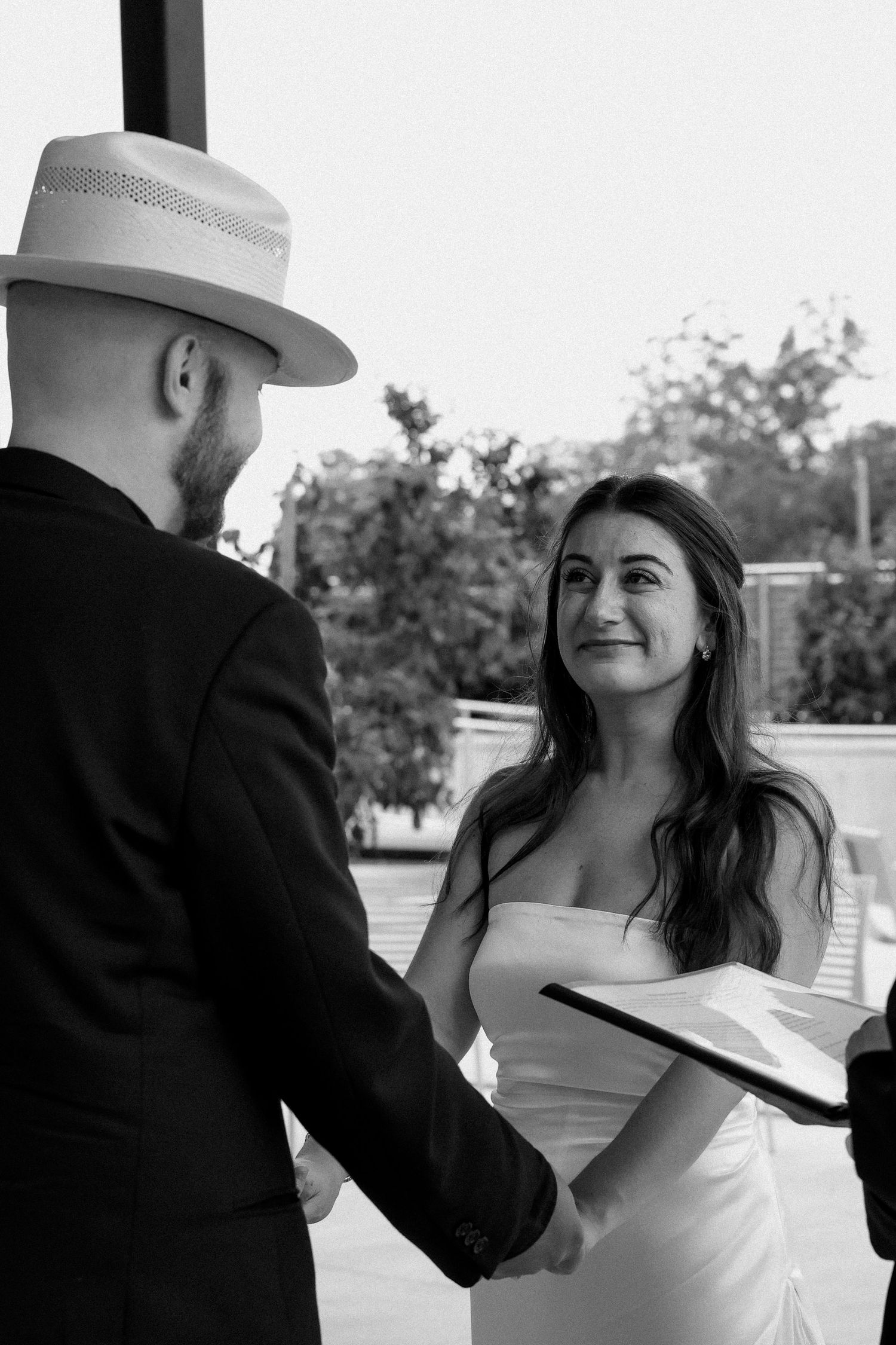 Black and white photo of bride gazing at groom in cowboy hat during outdoor wedding ceremony.
