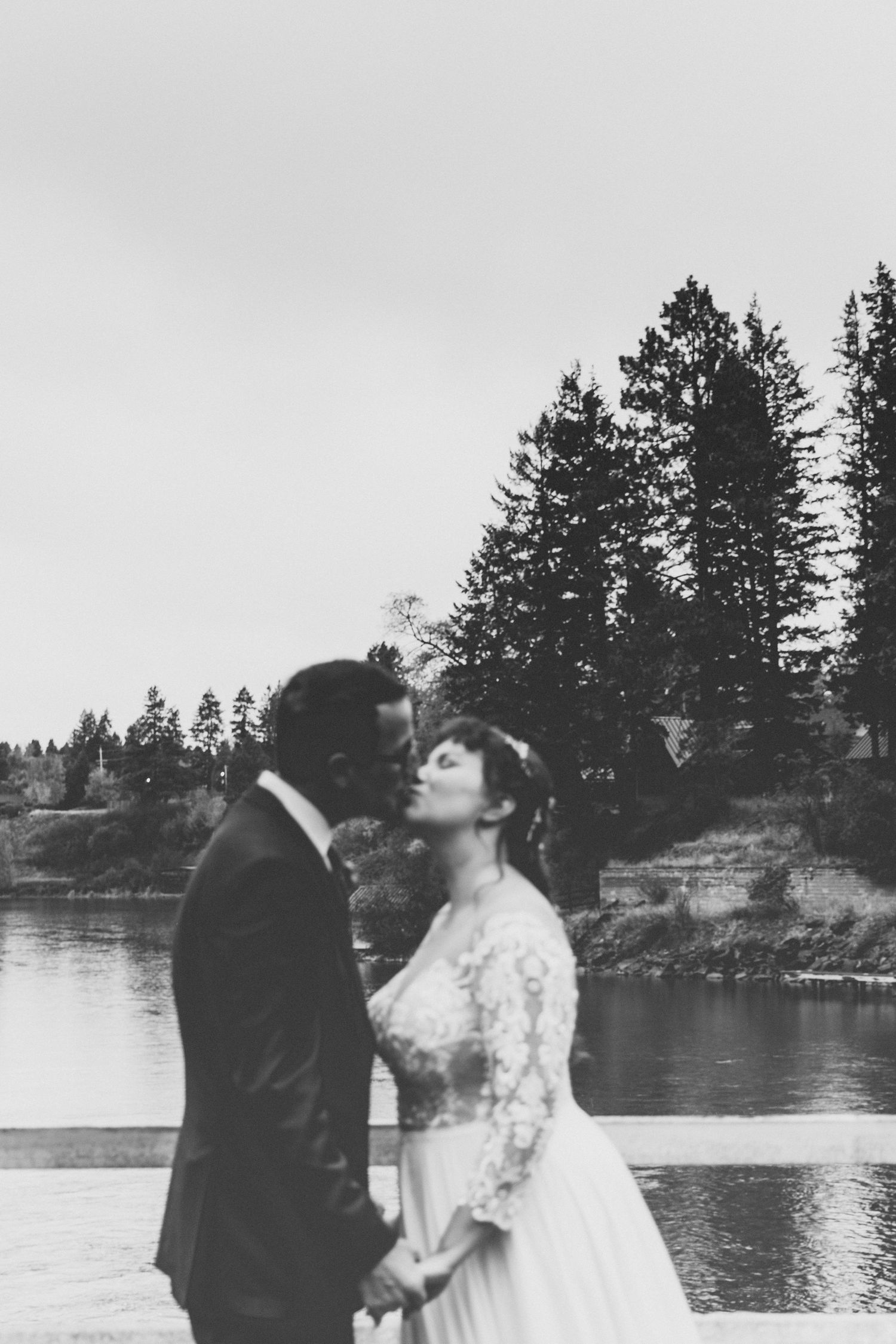 Black and white photo of bride and groom sharing a kiss by a serene lake with tall pine trees.
