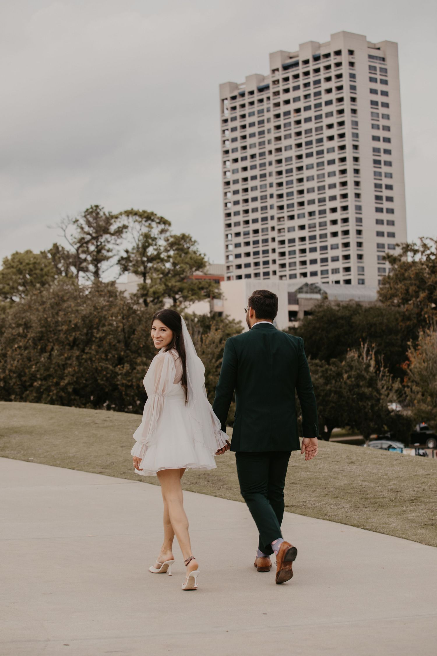 Bride in short white dress and groom in dark suit walk hand in hand near urban park.