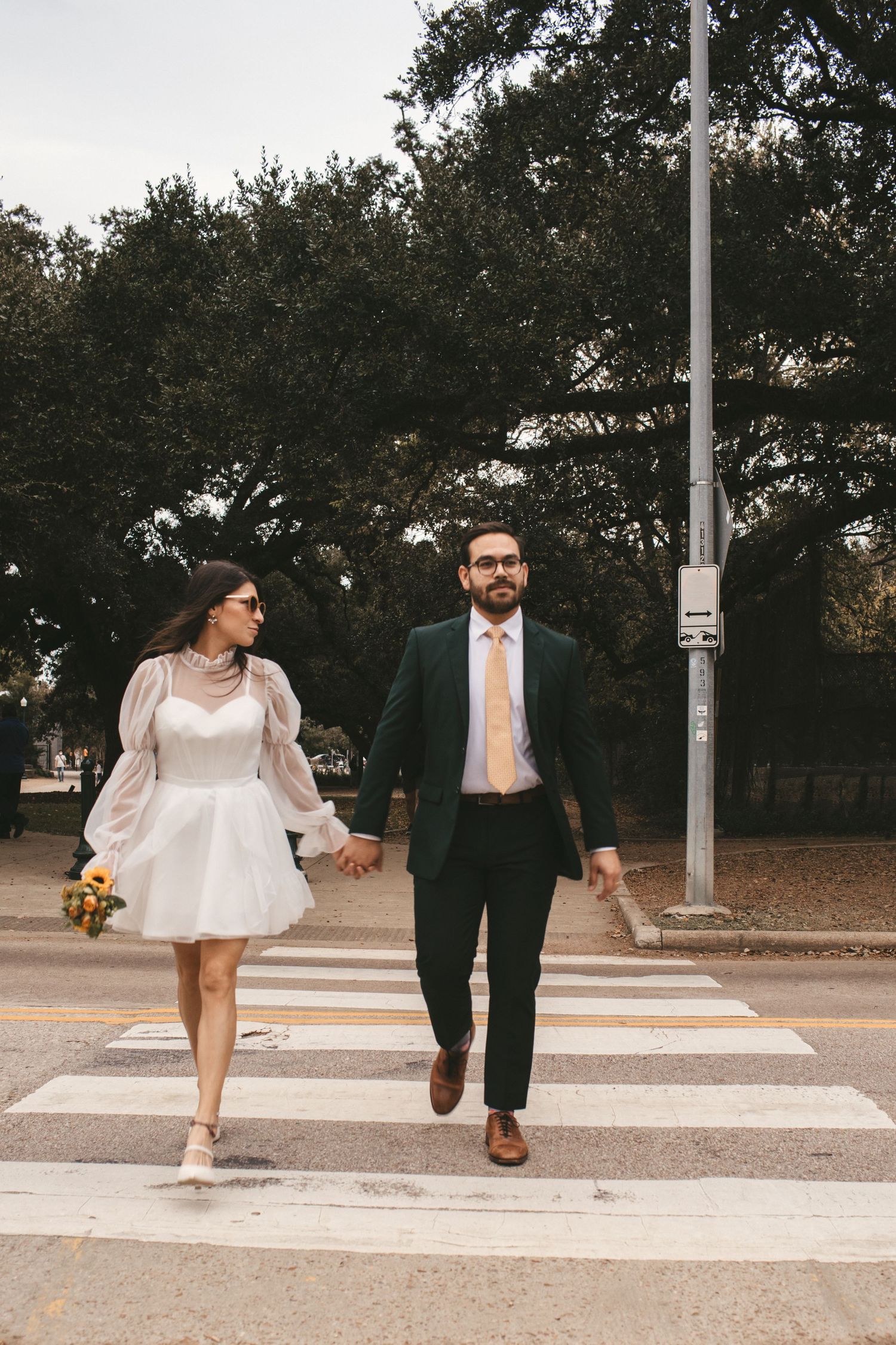 Bride and groom walk hand in hand across a crosswalk, she in white mini dress holding a colorful bouquet.