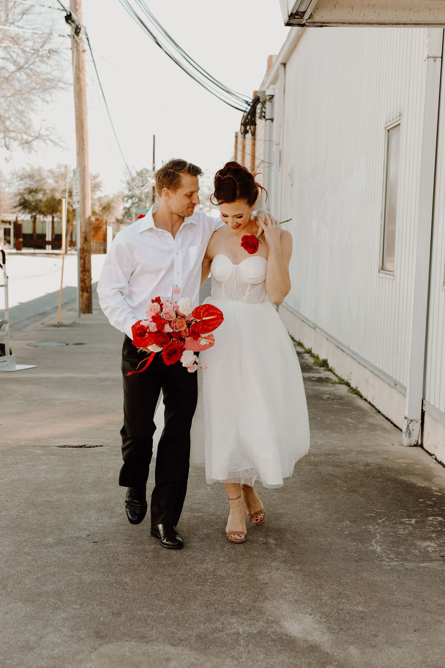 Bride in tea-length white dress and groom in white shirt walking together holding red floral bouquet.