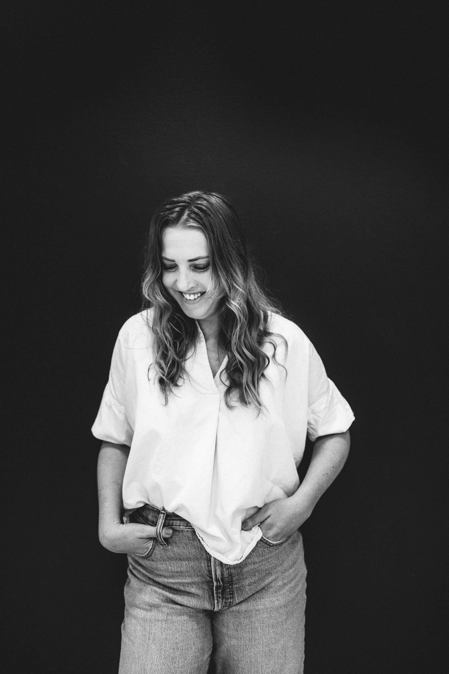 Black and white portrait of a smiling young woman in white top and jeans, hands in pockets.