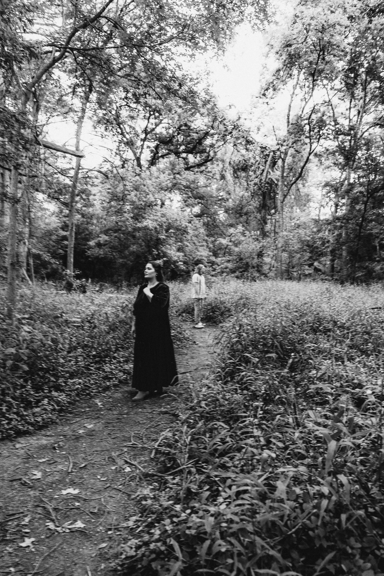 Black and white photo of a woman in a long dark dress standing on a forest path with another figure in the background.