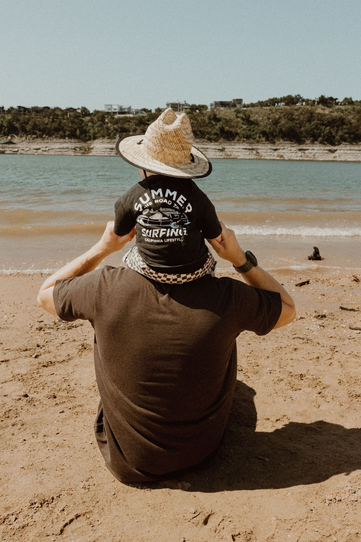 Father sitting on beach with baby in straw hat on his shoulders gazing at the water together.
