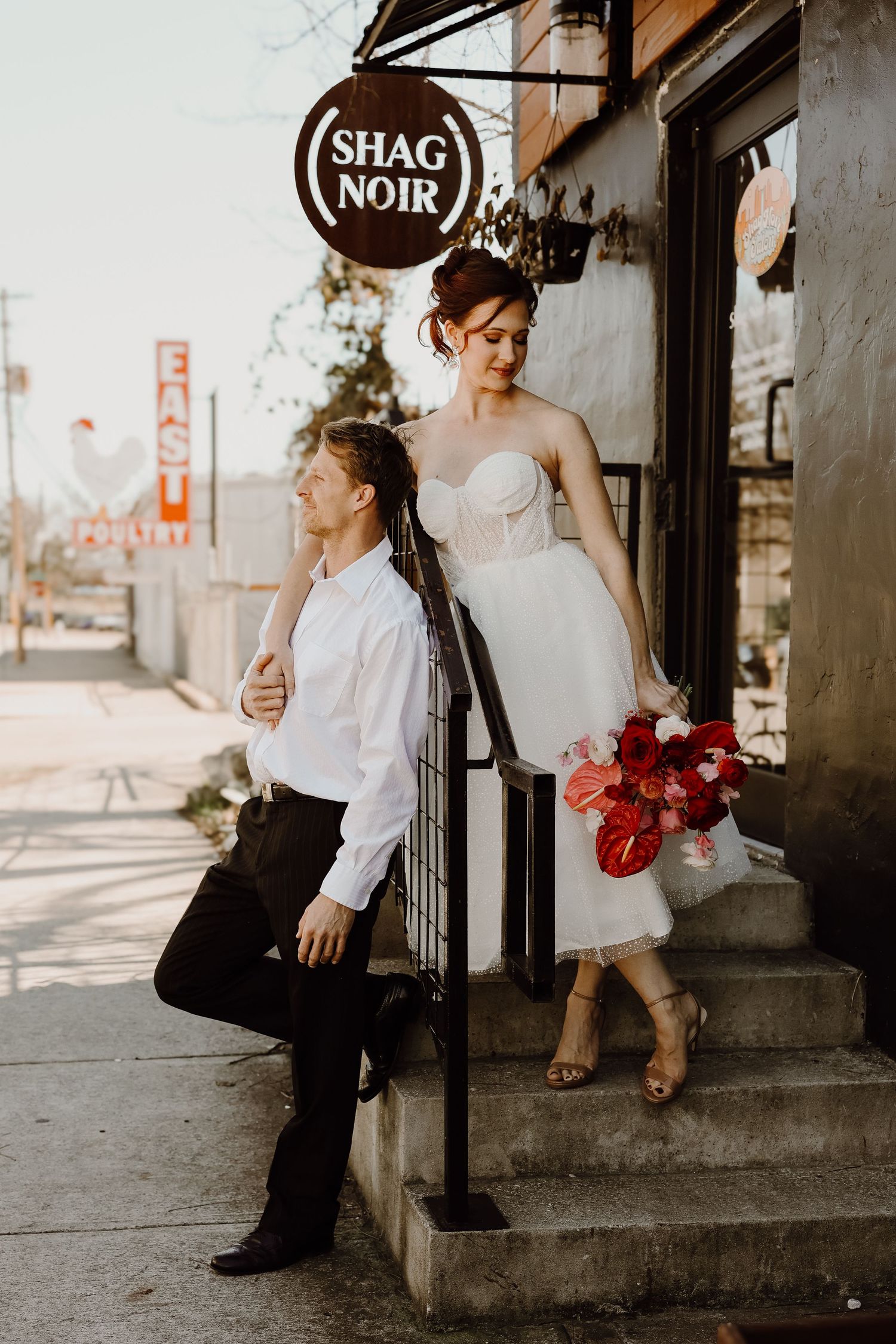 Bride in short tulle dress holding red bouquet poses with groom outside Shag Noir storefront.