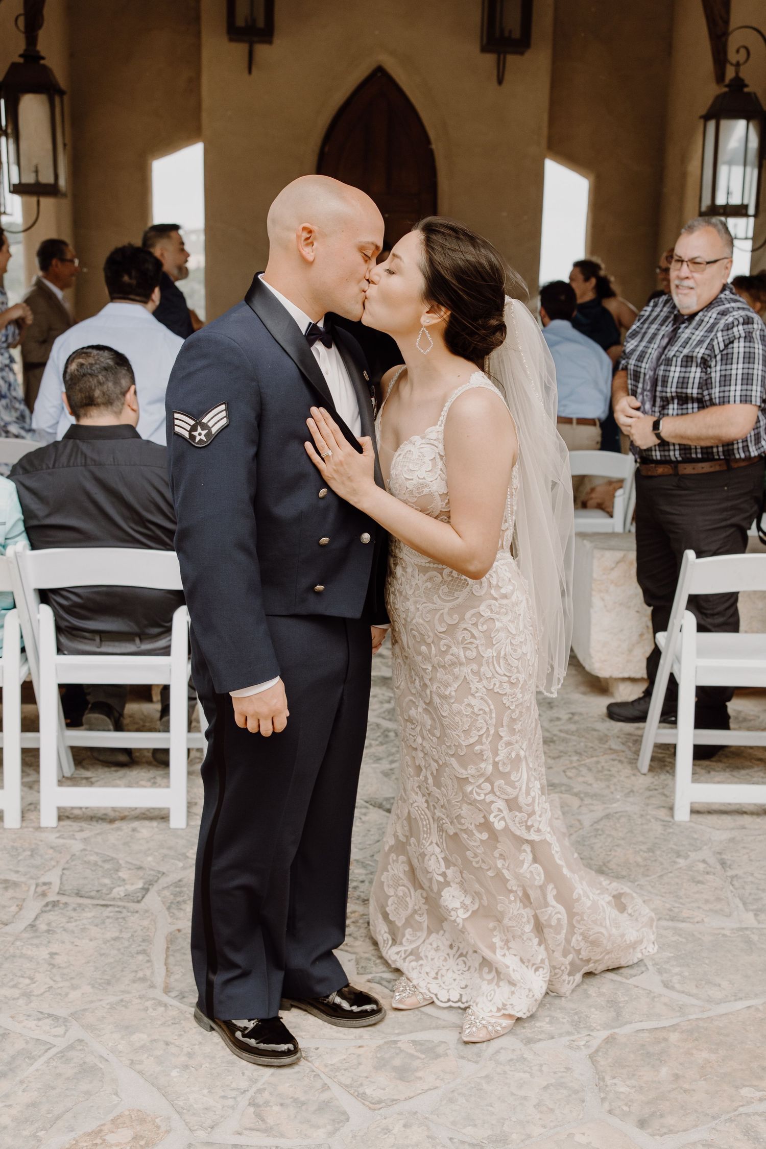 Military groom in dress uniform shares first kiss with bride in lace wedding gown at elegant chapel ceremony.