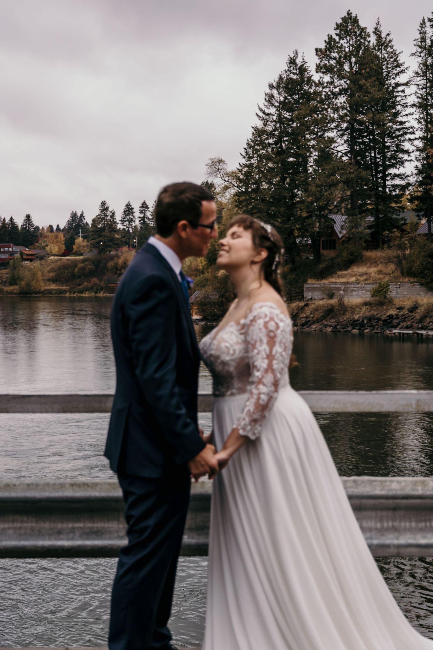 Bride and groom hold hands and lean in for a kiss on a bridge overlooking a misty autumn lake.
