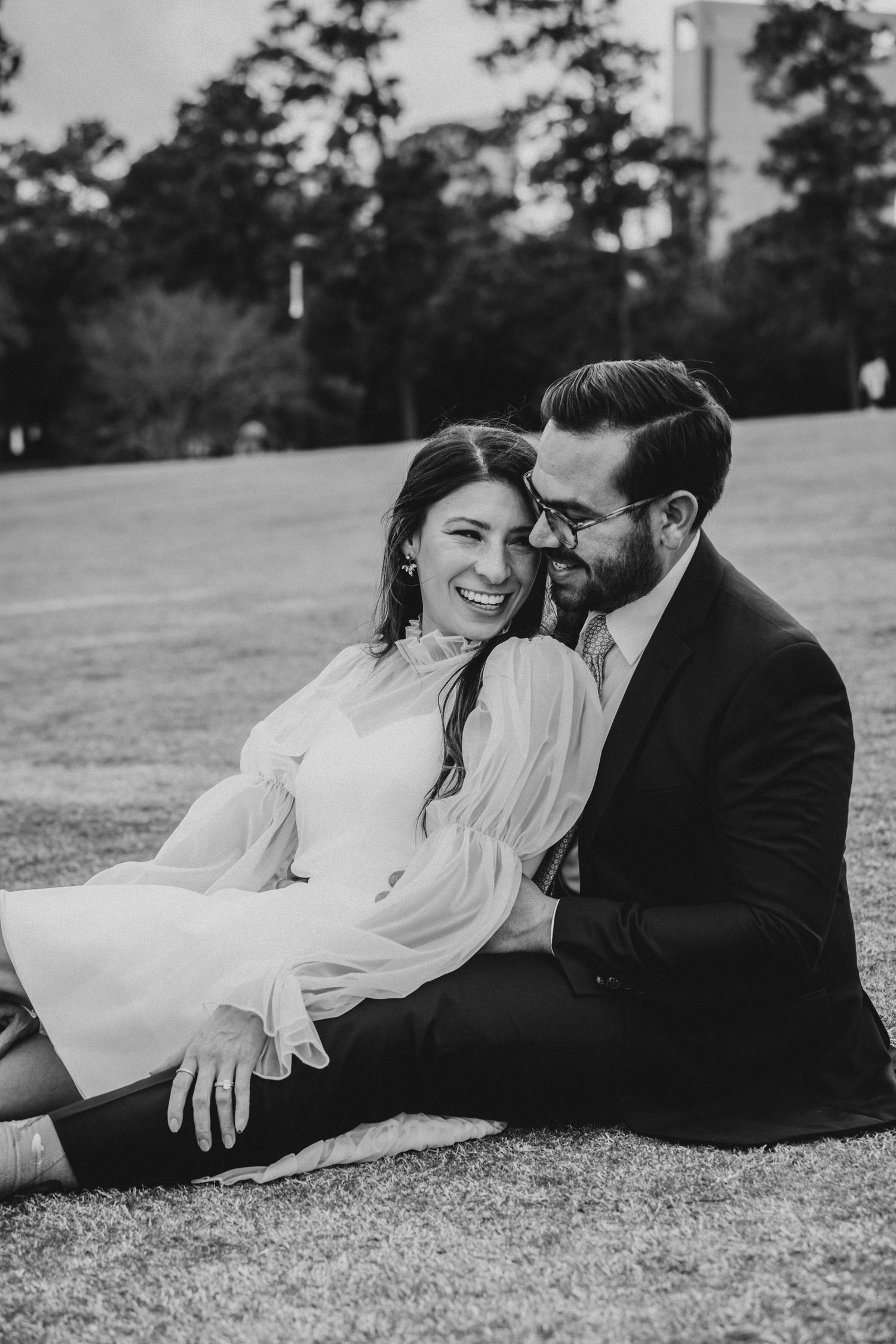 Black and white photo of laughing bride and groom sitting together on grass at park.
