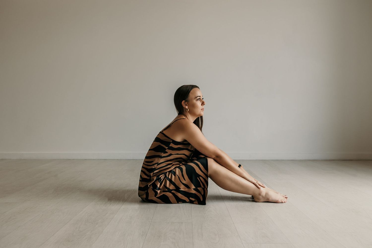Woman in a tiger print satin dress sitting barefoot on a light wood studio floor.