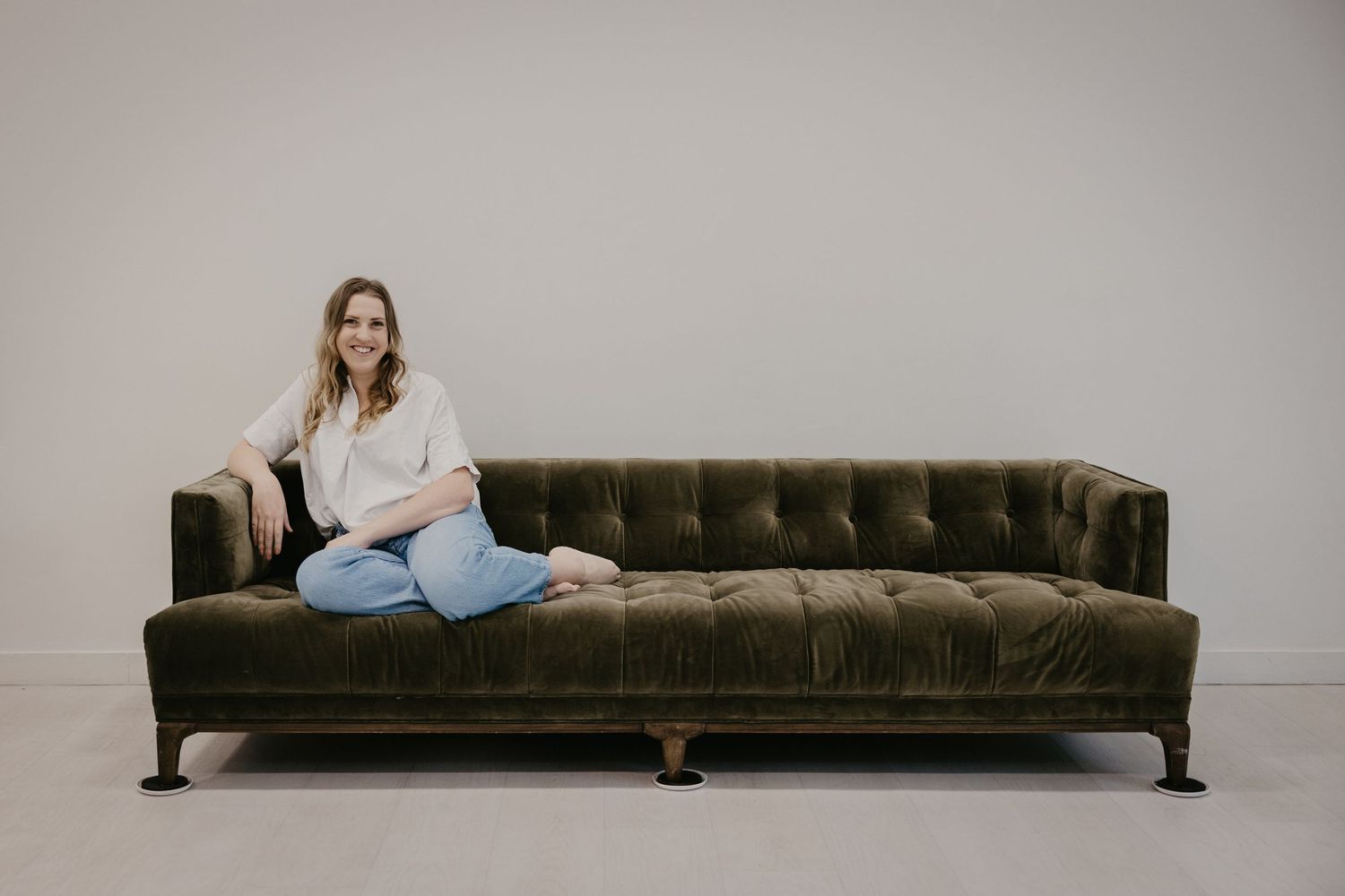 Smiling woman in white blouse and jeans sitting relaxed on a green tufted velvet sofa.