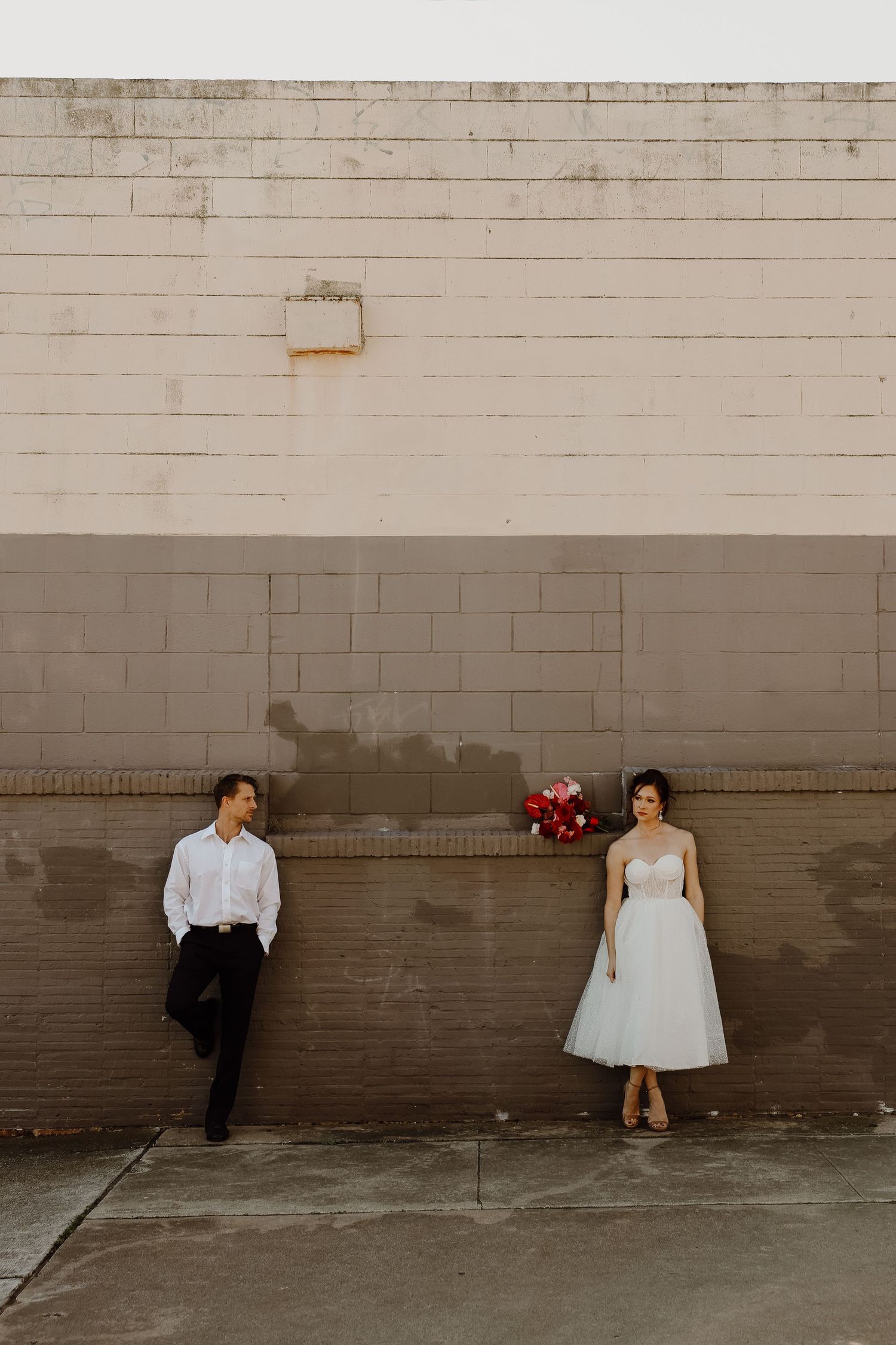 Bride in tea-length tulle dress and groom stand apart against a large concrete wall with red bouquet.
