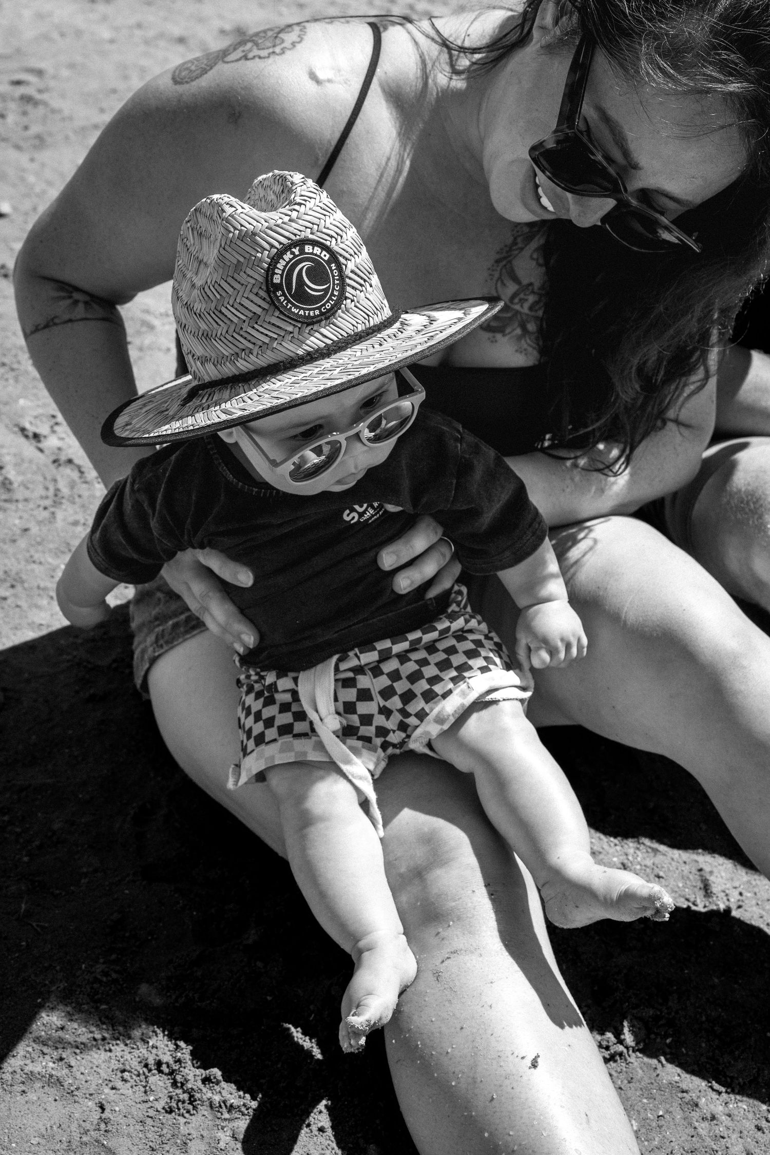 Black and white photo of baby wearing straw hat and sunglasses sitting on mothers lap at beach.