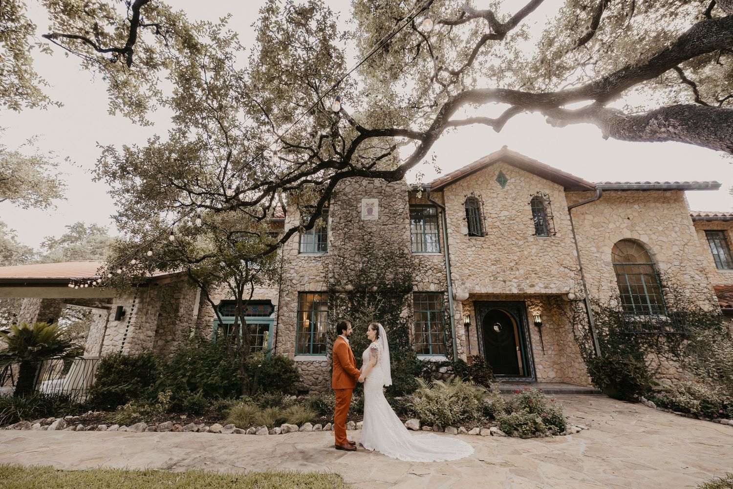 Couple in wedding attire standing before a rustic stone mansion surrounded by large oak trees.