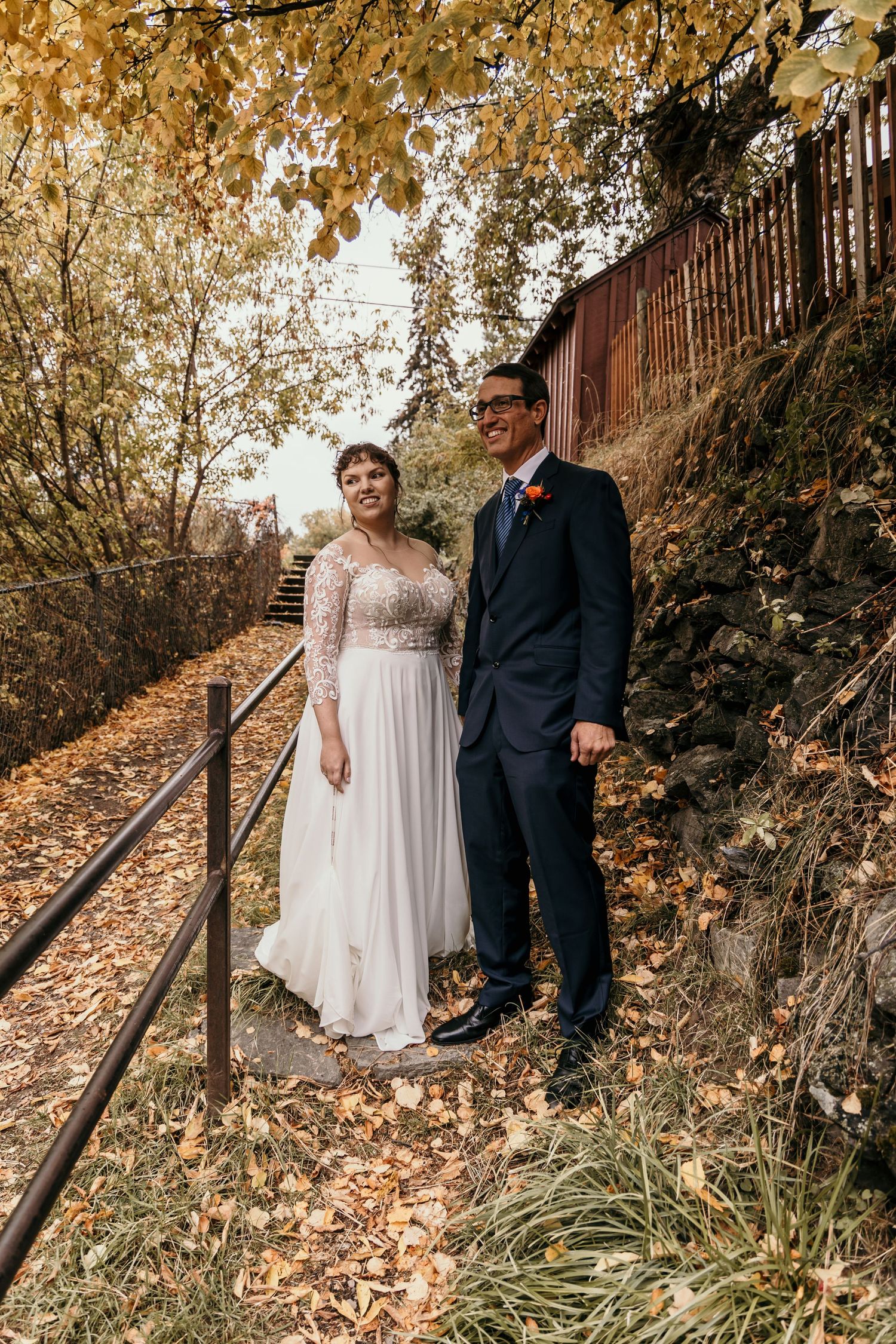 Smiling bride and groom stand together on a leaf-covered path surrounded by golden autumn foliage.