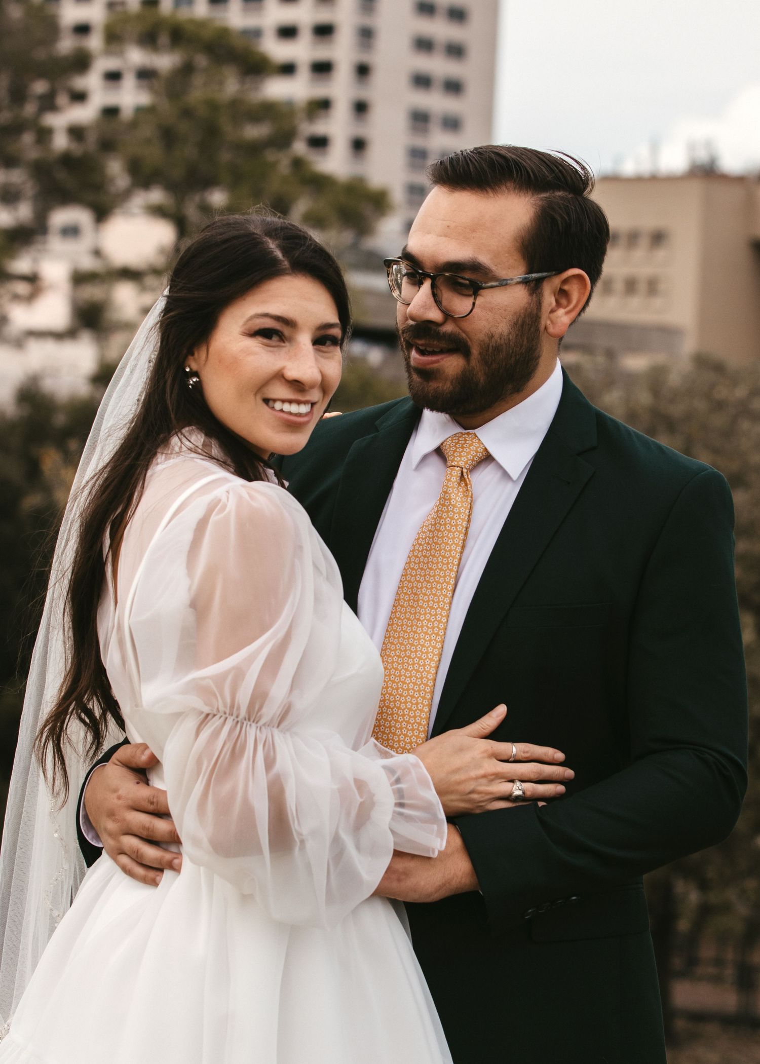 Smiling bride in sheer white gown embraces groom in dark suit with gold tie on rooftop.
