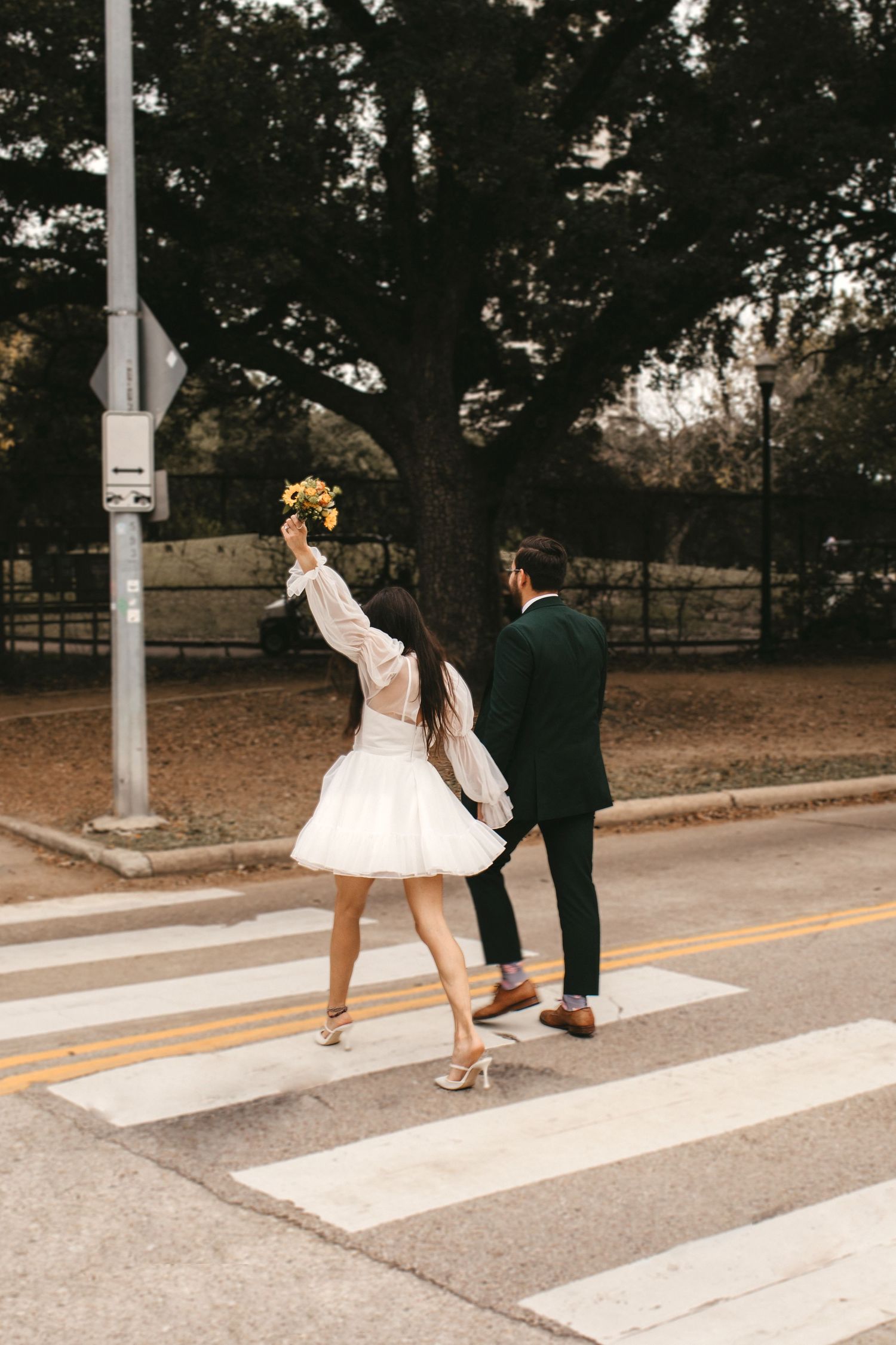 Bride raises yellow bouquet while crossing street with groom in dark suit on a zebra crossing.