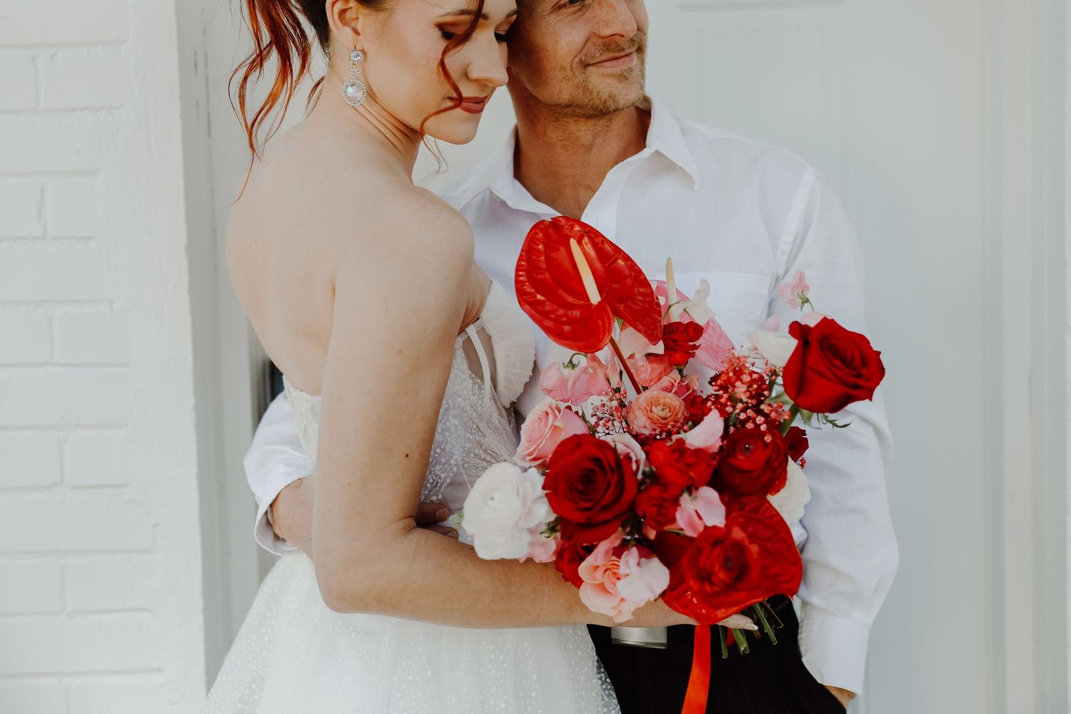 Bride and groom embracing tenderly, bride holding a vibrant red and pink rose wedding bouquet.