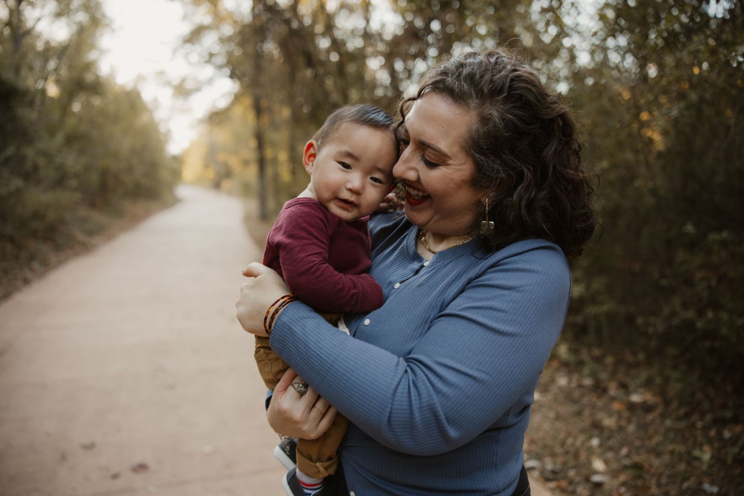 A woman tenderly holds a smiling baby on a tree-lined path during an outdoor family photo session.