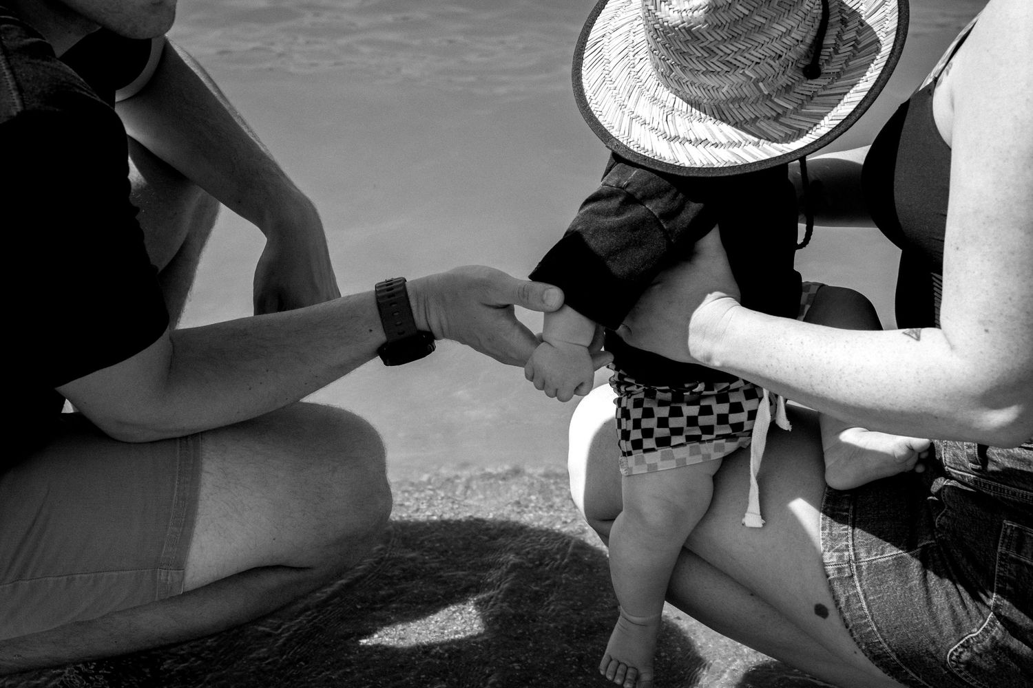 Black and white close-up of parents holding their baby's hands near the water's edge at the beach.