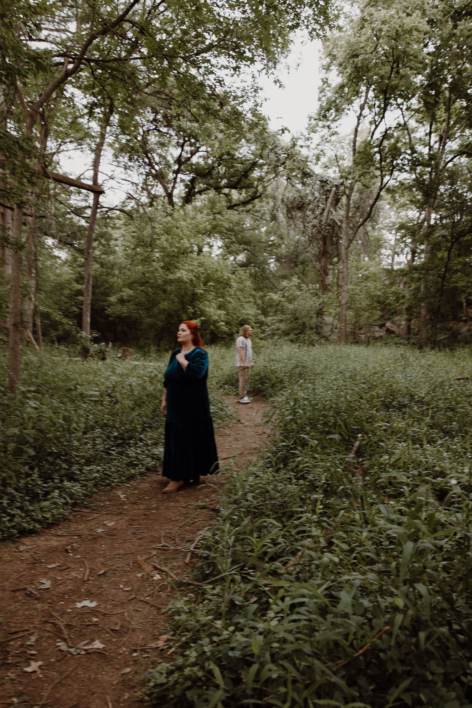 A woman with red hair in a black maxi dress stands on a woodland path surrounded by lush greenery with another person behind her.