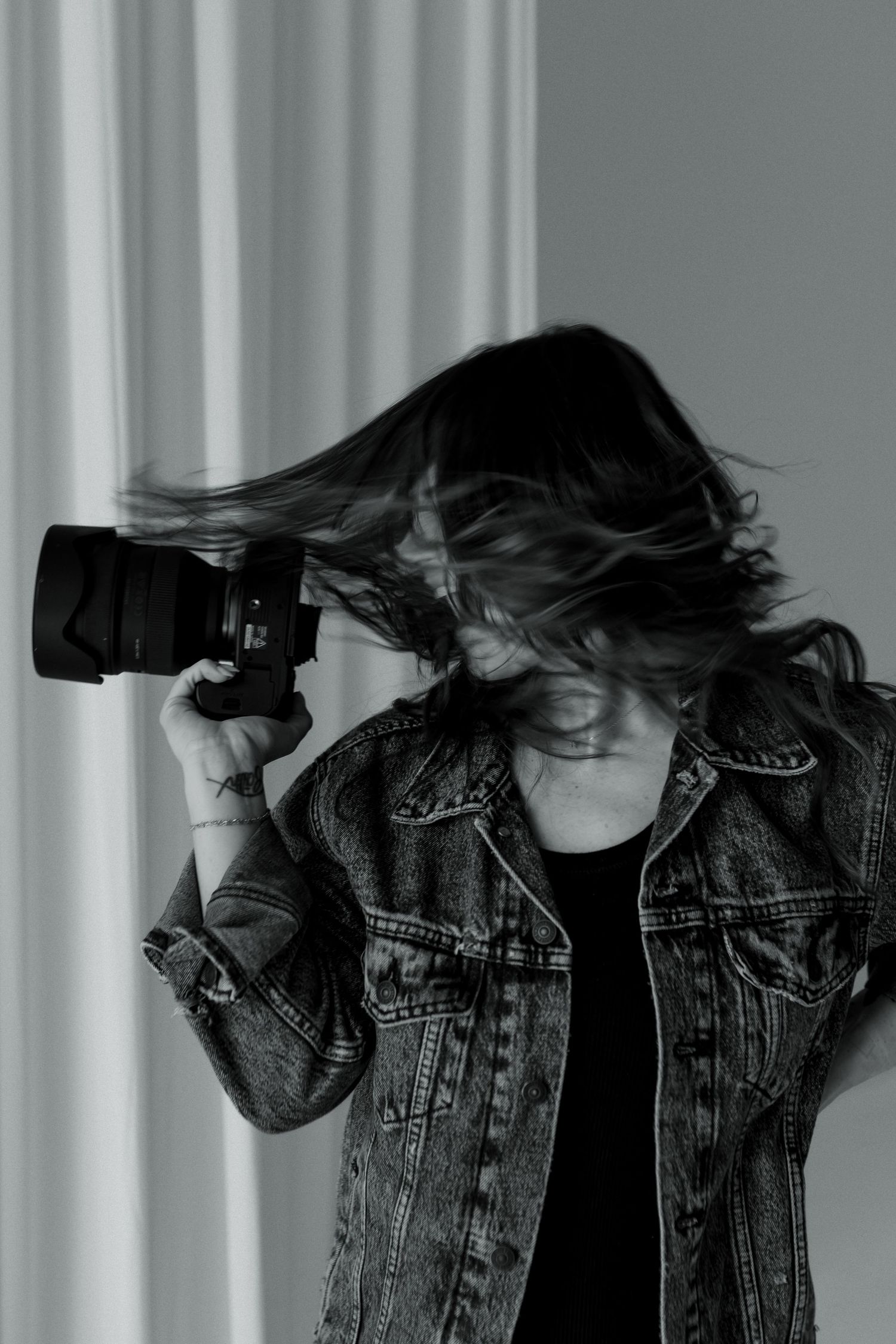 Photographer in denim jacket with hair blowing, holding camera up in dramatic black and white portrait.