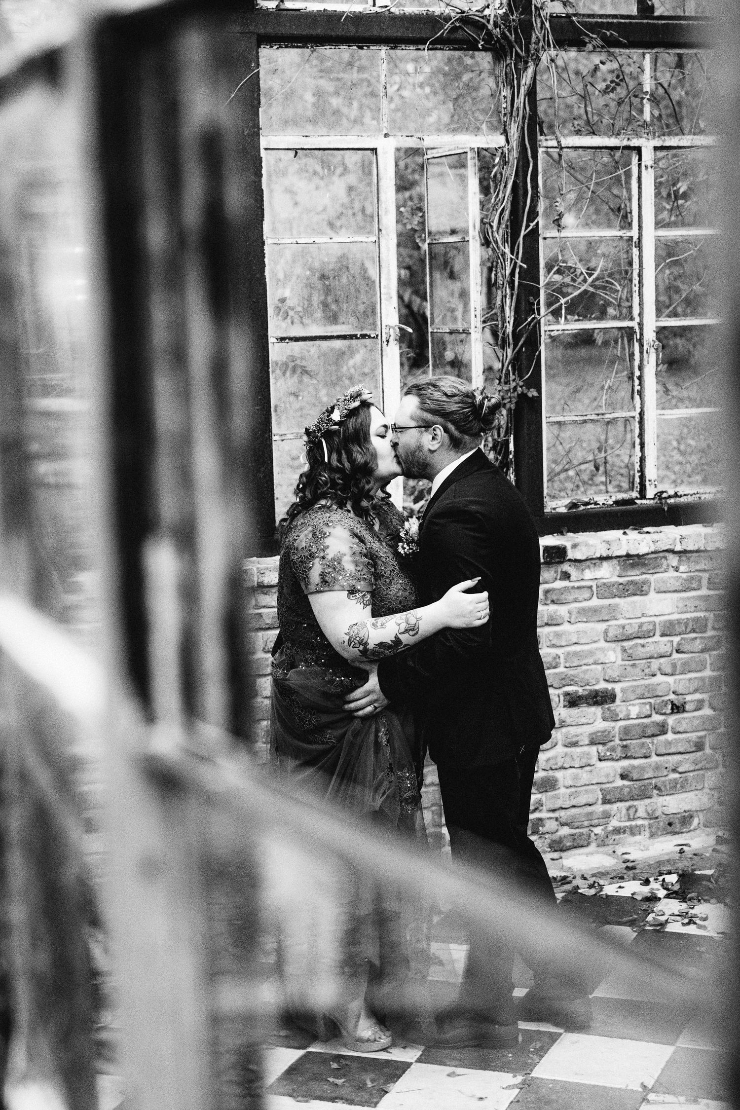 Black and white photo of bride and groom sharing a kiss in rustic brick greenhouse.
