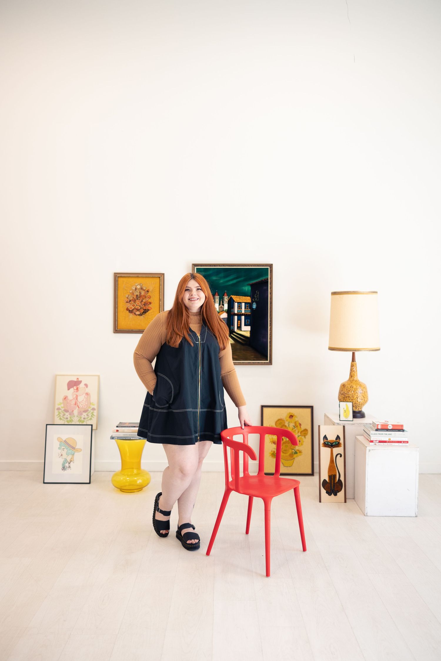 Smiling redheaded woman in navy dress standing beside red chair among vintage art collection.