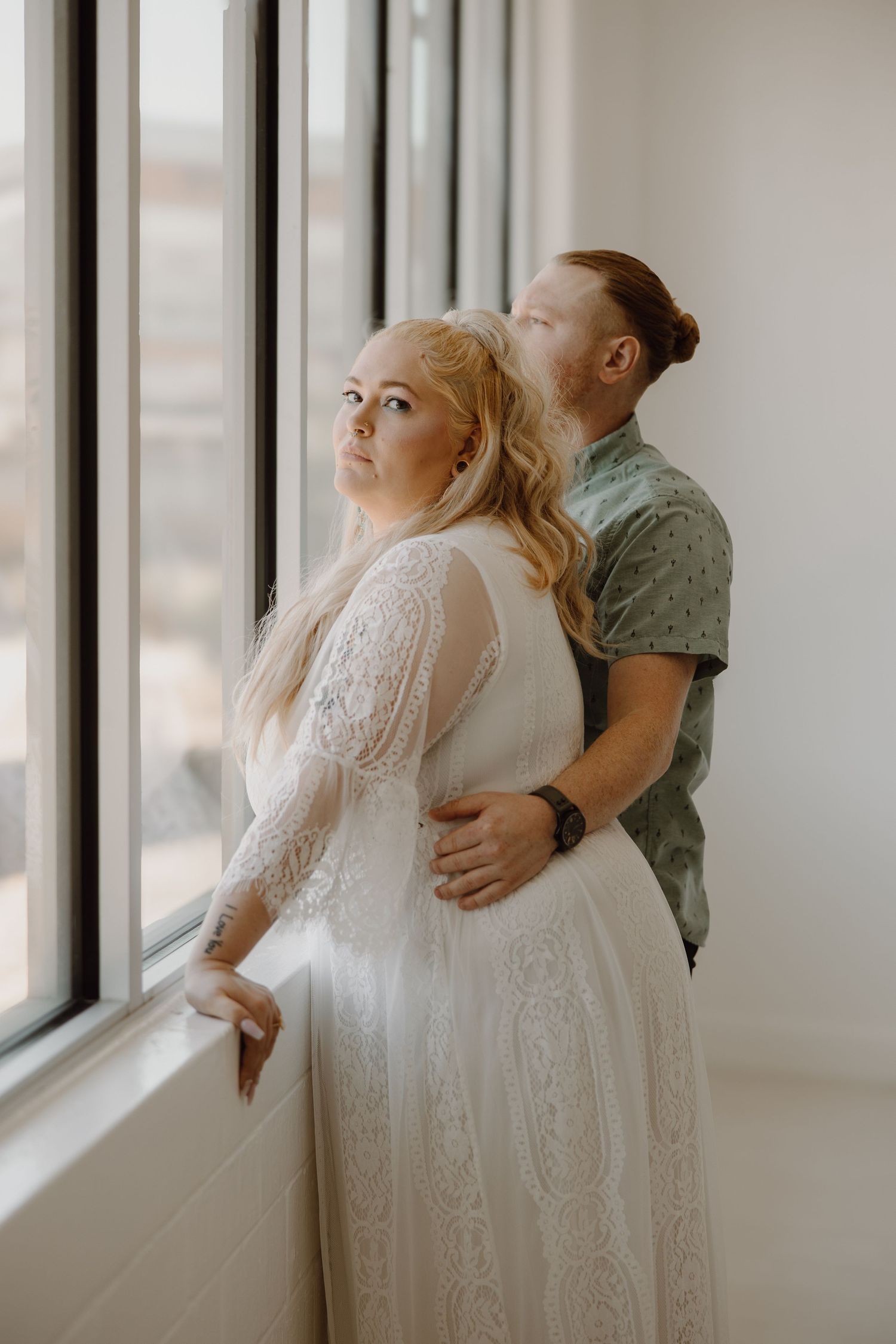 Groom embracing bride from behind near large window, both gazing upward in soft light.