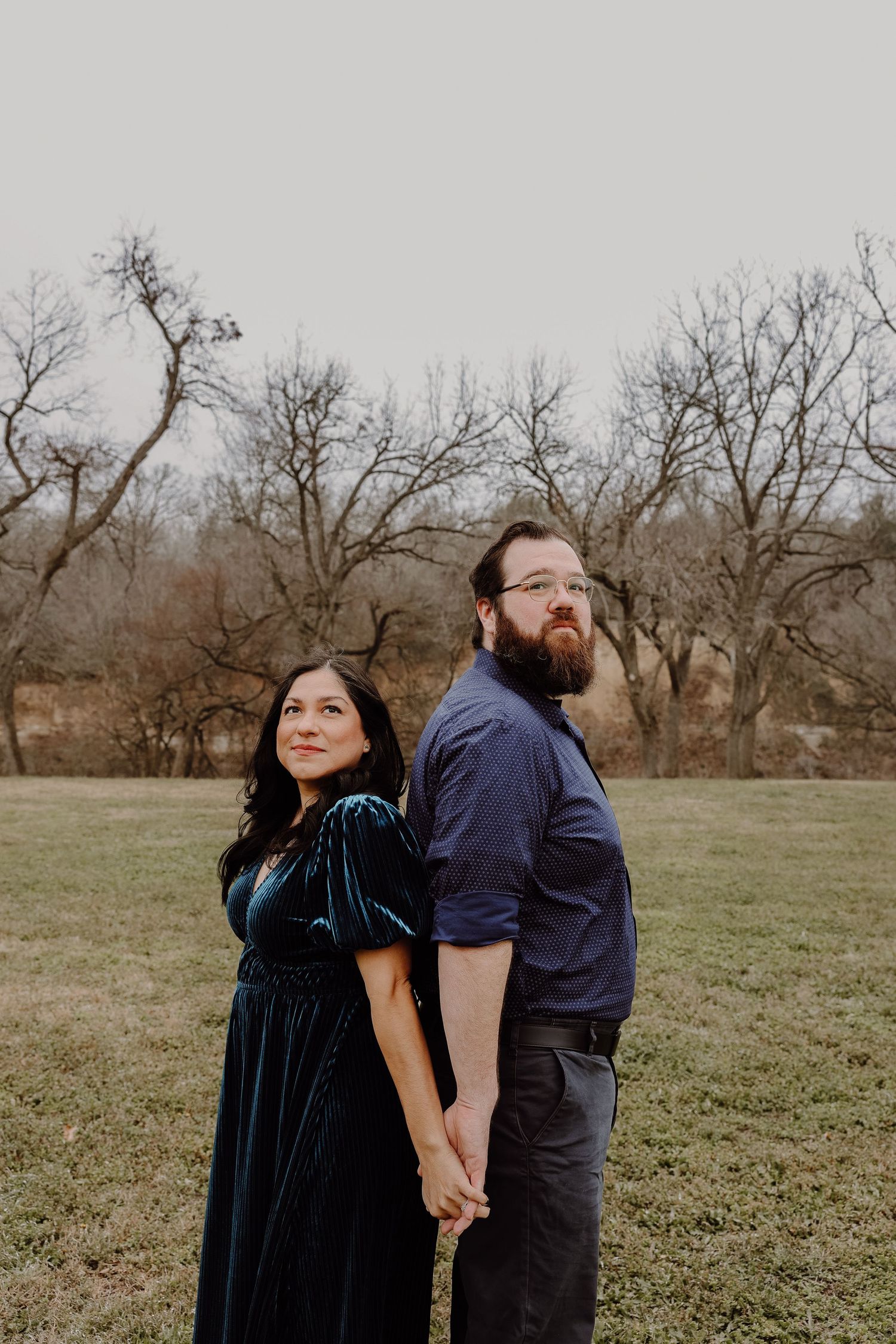 Couple holding hands back to back in field, woman in teal velvet dress with bare trees behind.