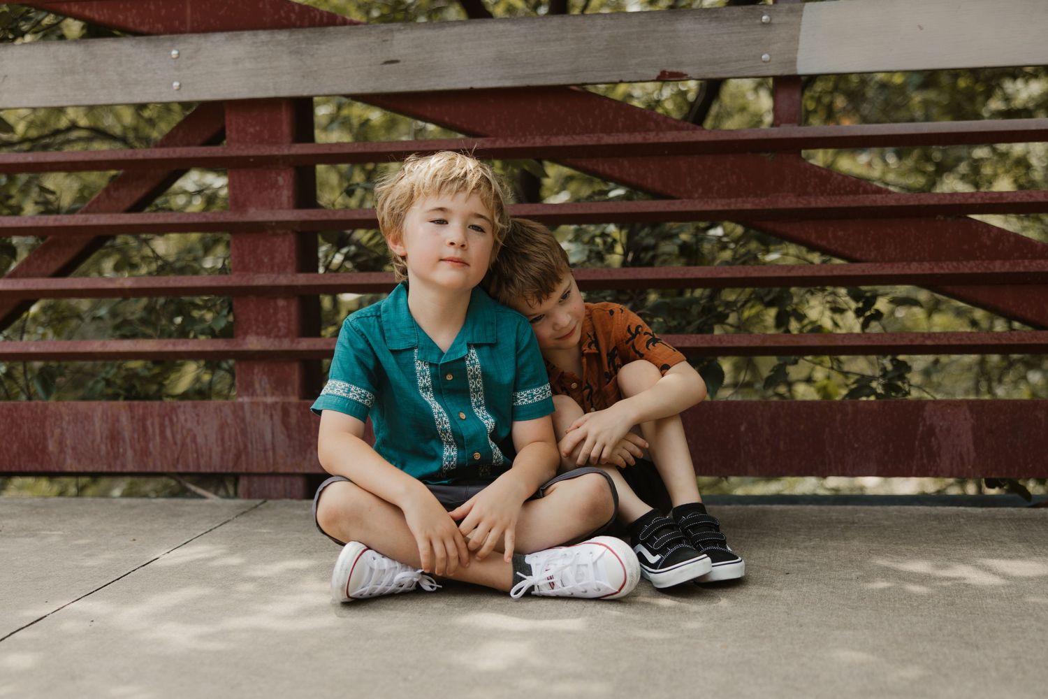 Two young blonde brothers sit together on a bridge in a candid outdoor portrait photo.
