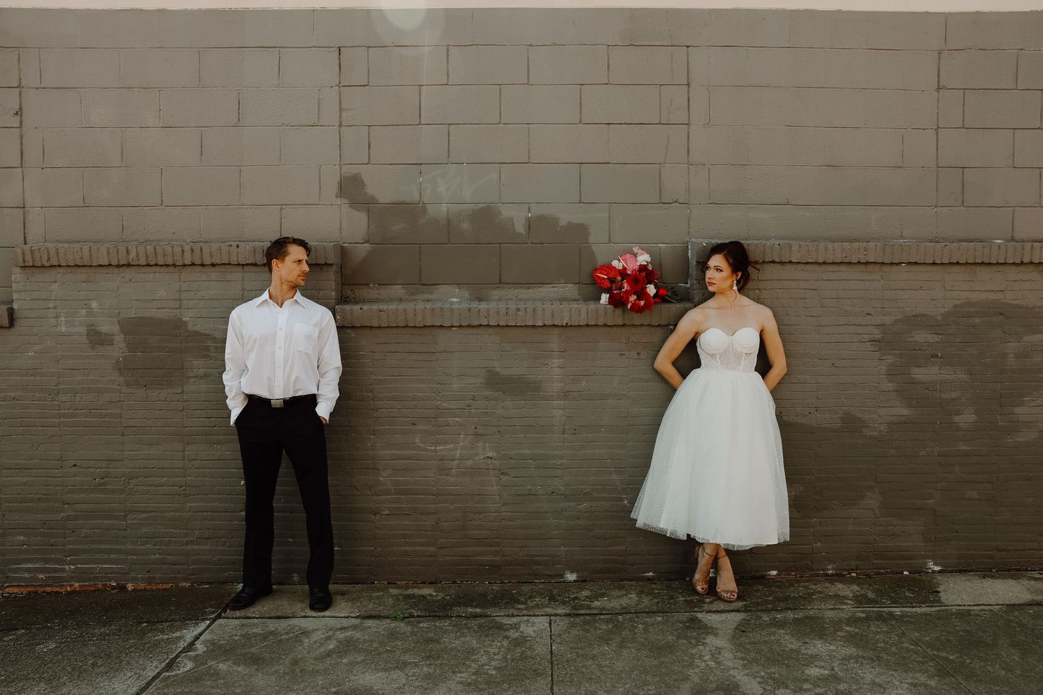 Bride in tea-length white dress holds red bouquet while groom stands apart against urban gray brick wall.