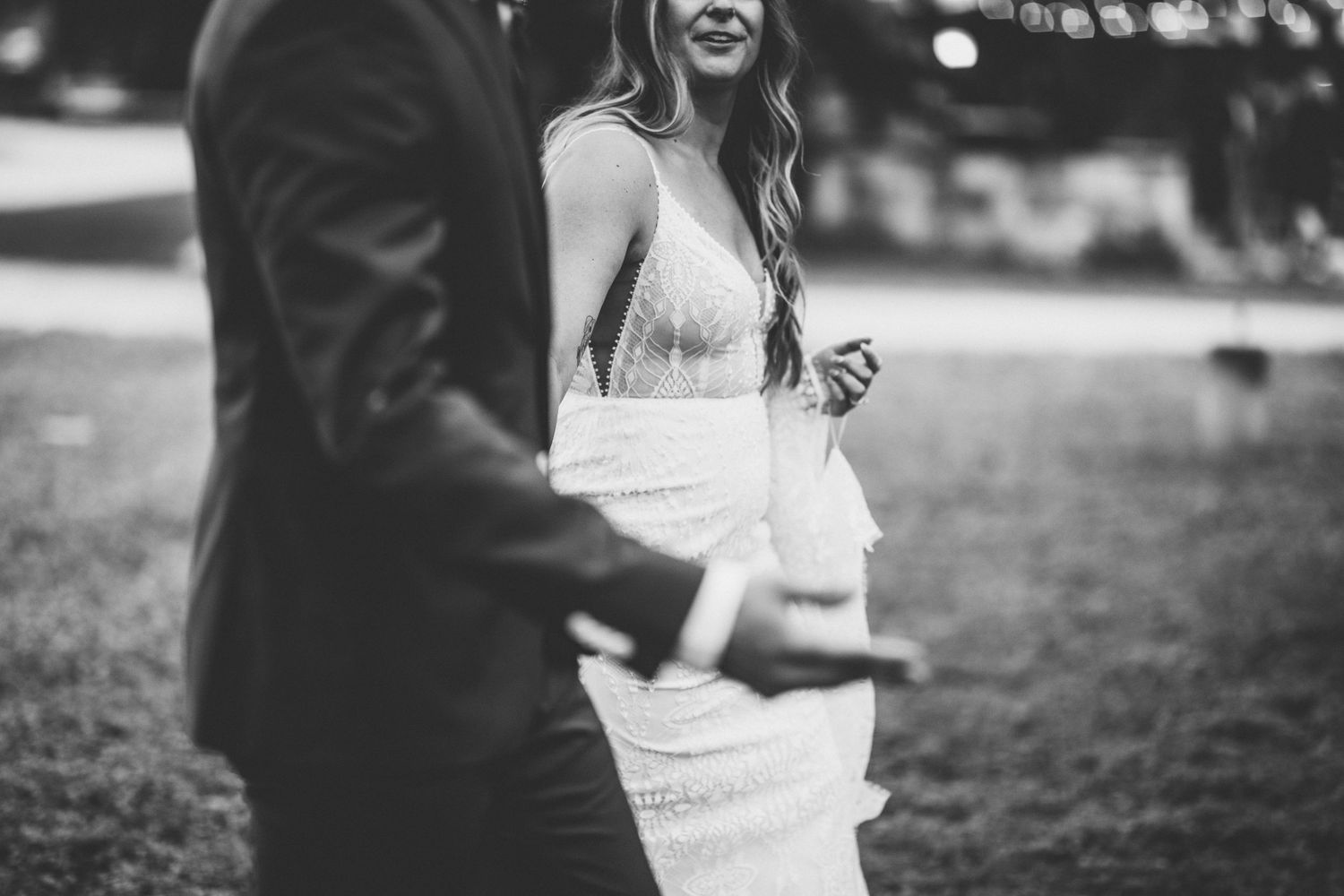 Black and white photo of bride in backless lace gown holding grooms hand while smiling outdoors.