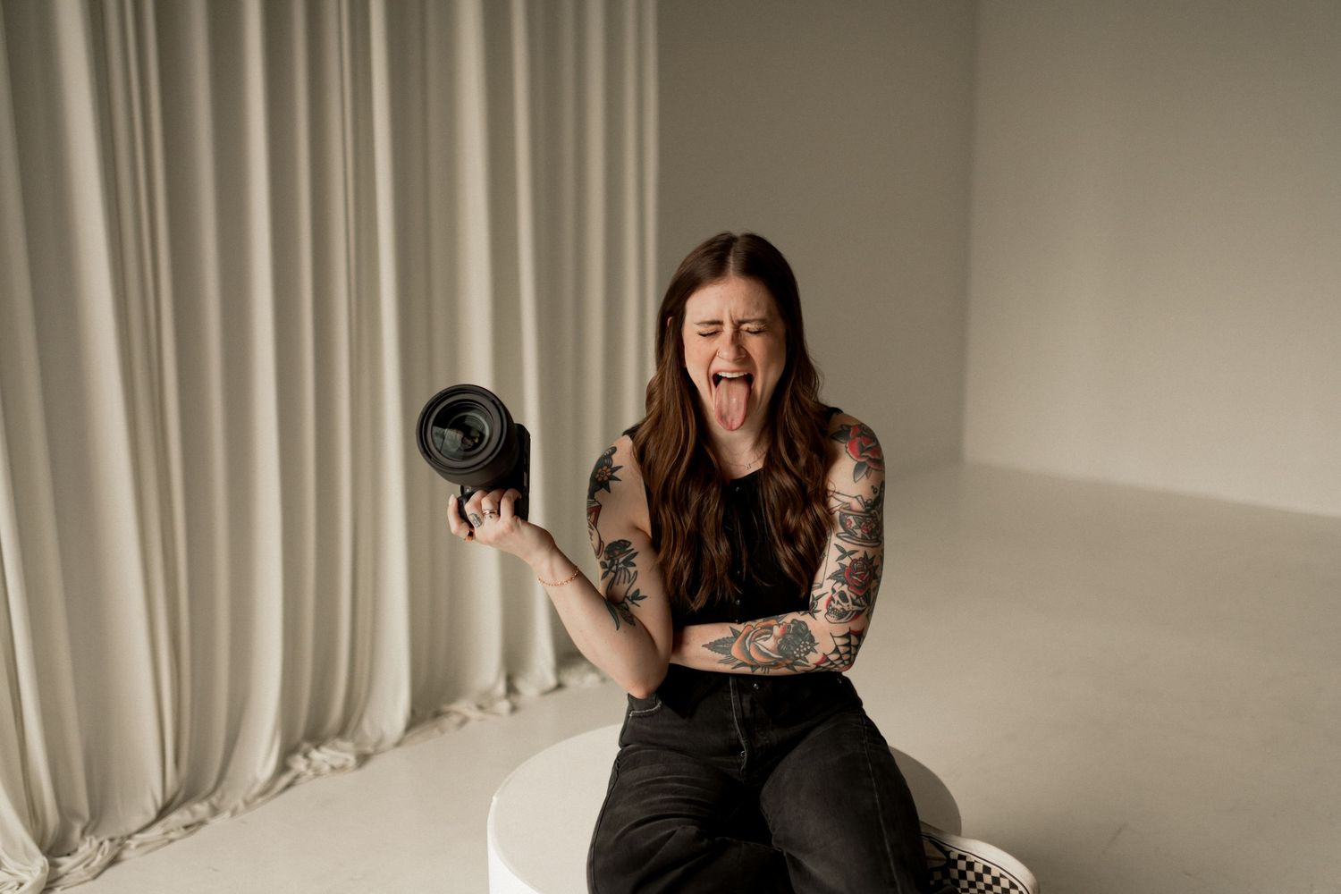 Tattooed woman sitting on white pedestal holding camera lens, laughing expressively against neutral curtain backdrop.